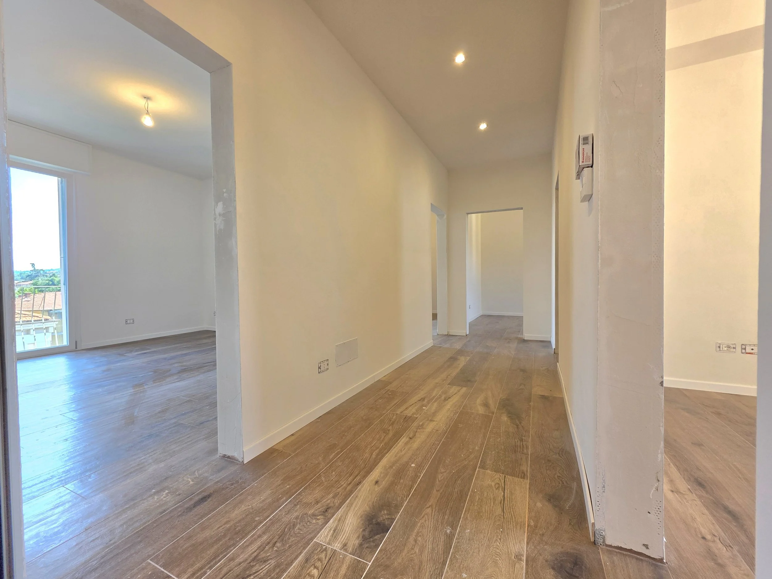 Interno di un appartamento nuovo con pareti bianche, pavimento in gres effetto legno, porte nuove, vista su vari ambienti e una finestra con balcone di fronte che lascia entrare luce naturale.
