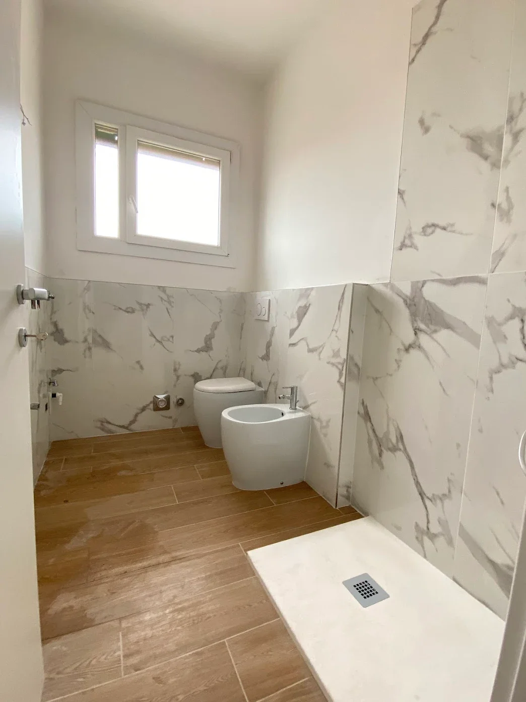 Bagno con pareti in marmo bianco e piastrelle in legno, water e bidet, finestra sulla parete superiore