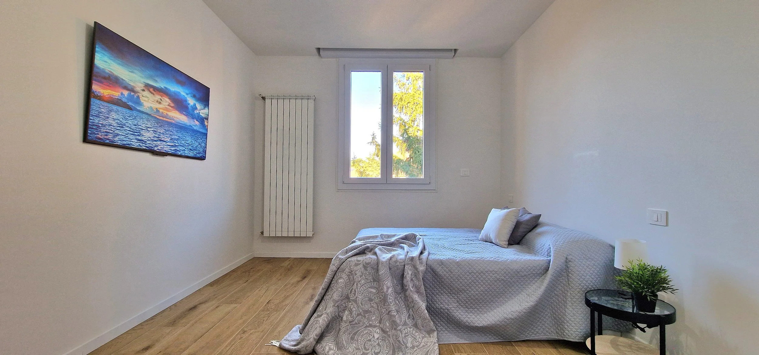 Camera da letto minimalista con letto, due cuscini, coperta, finestra, radiatore e quadro con paesaggio marino al tramonto sulla parete.