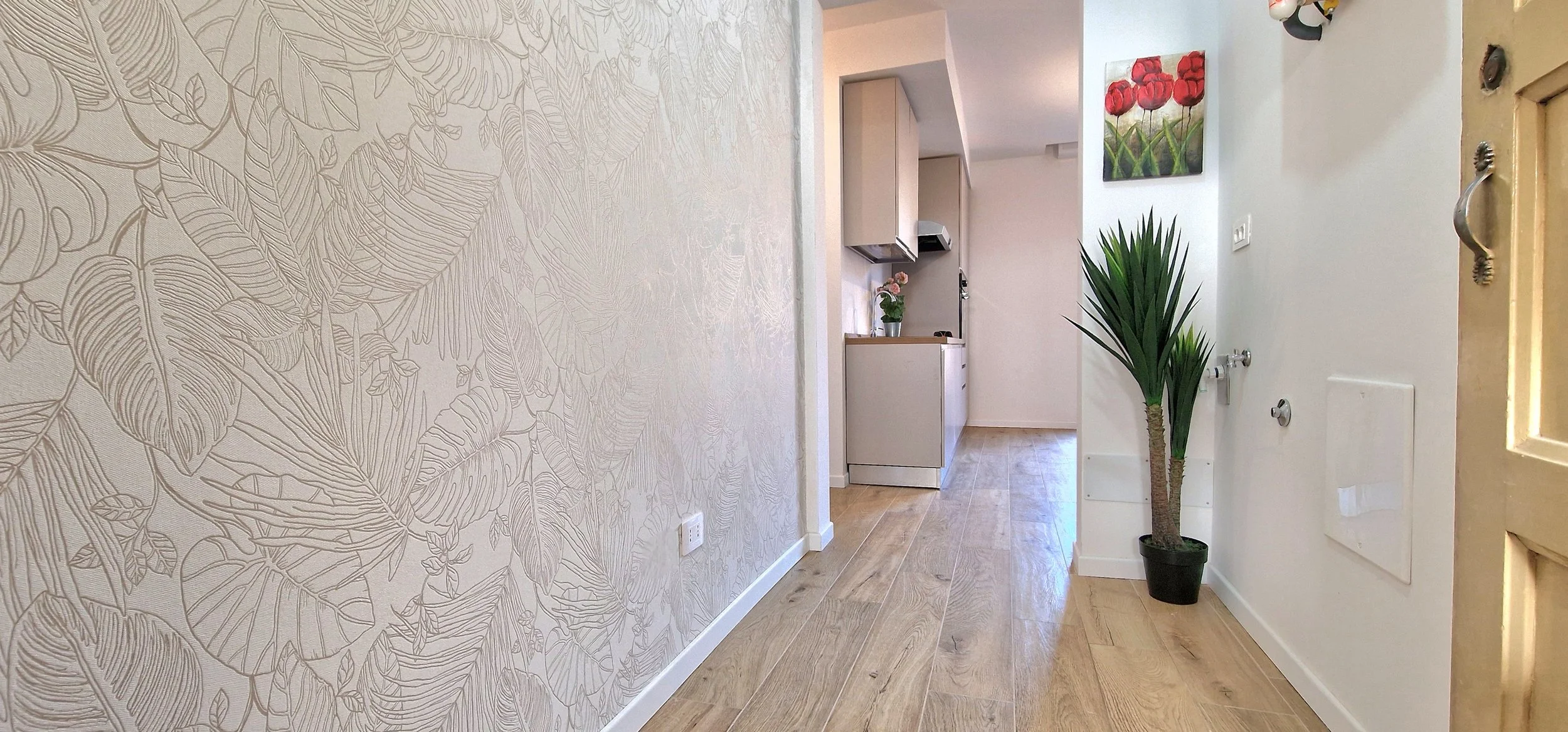 Interno di una casa con un corridoio, una parete con carta da parati a motivo di foglie, una pianta in un vaso nero e un quadro con fiori rossi sulla parete. Sul lato destro, una porta chiusa.
