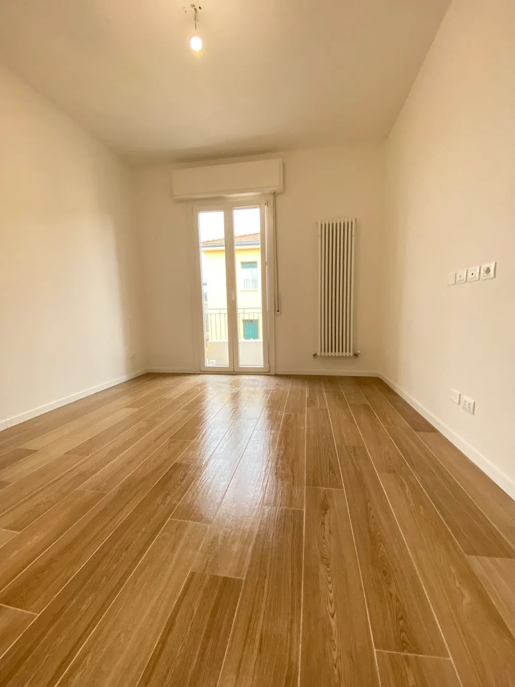 Stanza vuota con pavimento in legno e porta finestra che dà su un balcone. Pareti bianche, radiatore e presa elettrica visibili.