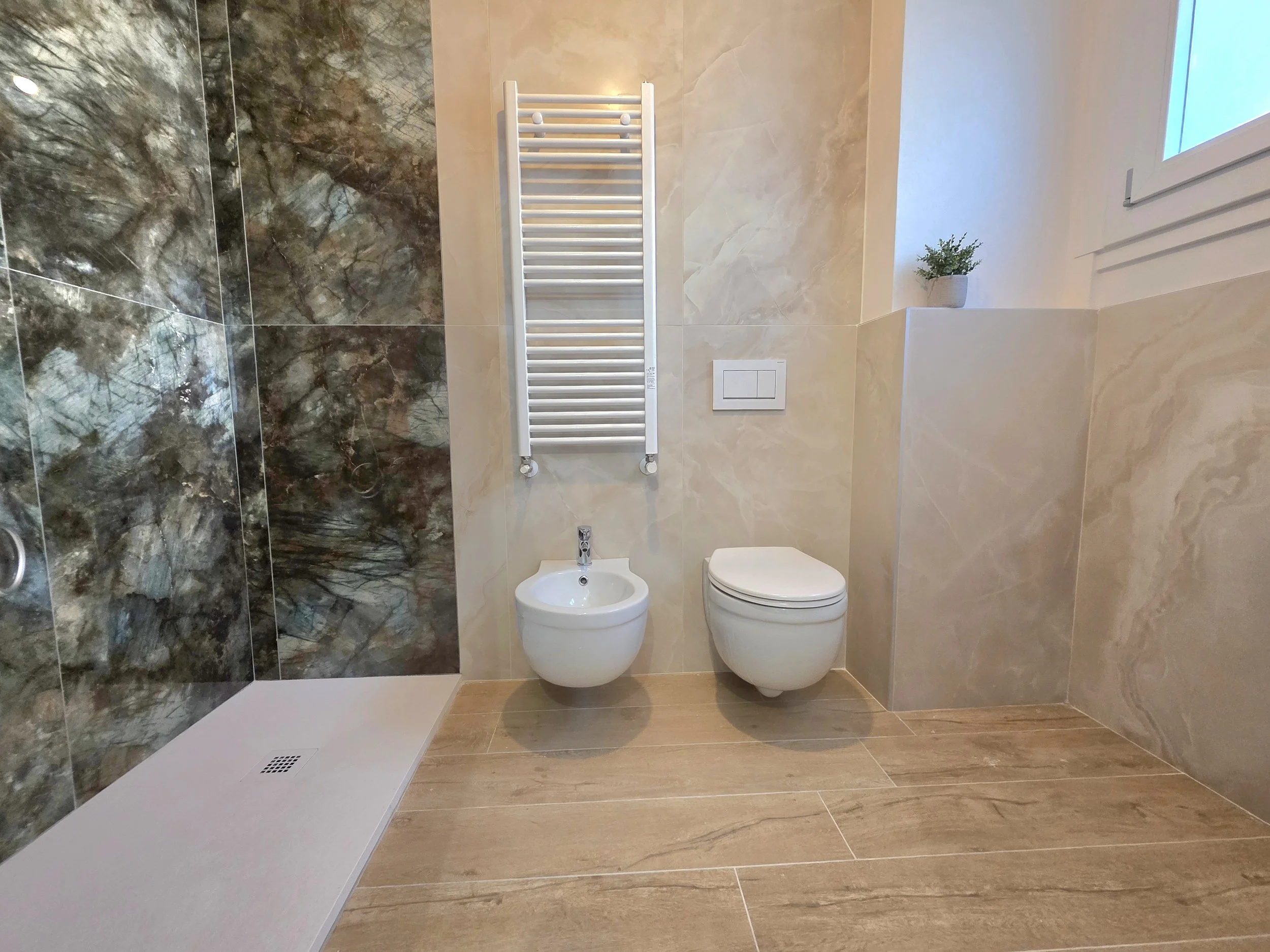 Bagno moderno con pareti rivestite in marmo beige e una parete con grande finestra, pavimento in piastrelle legno naturale, sanitari bianchi tra cui bidet, wc e termoscaldino bianco con barre orizzontali.