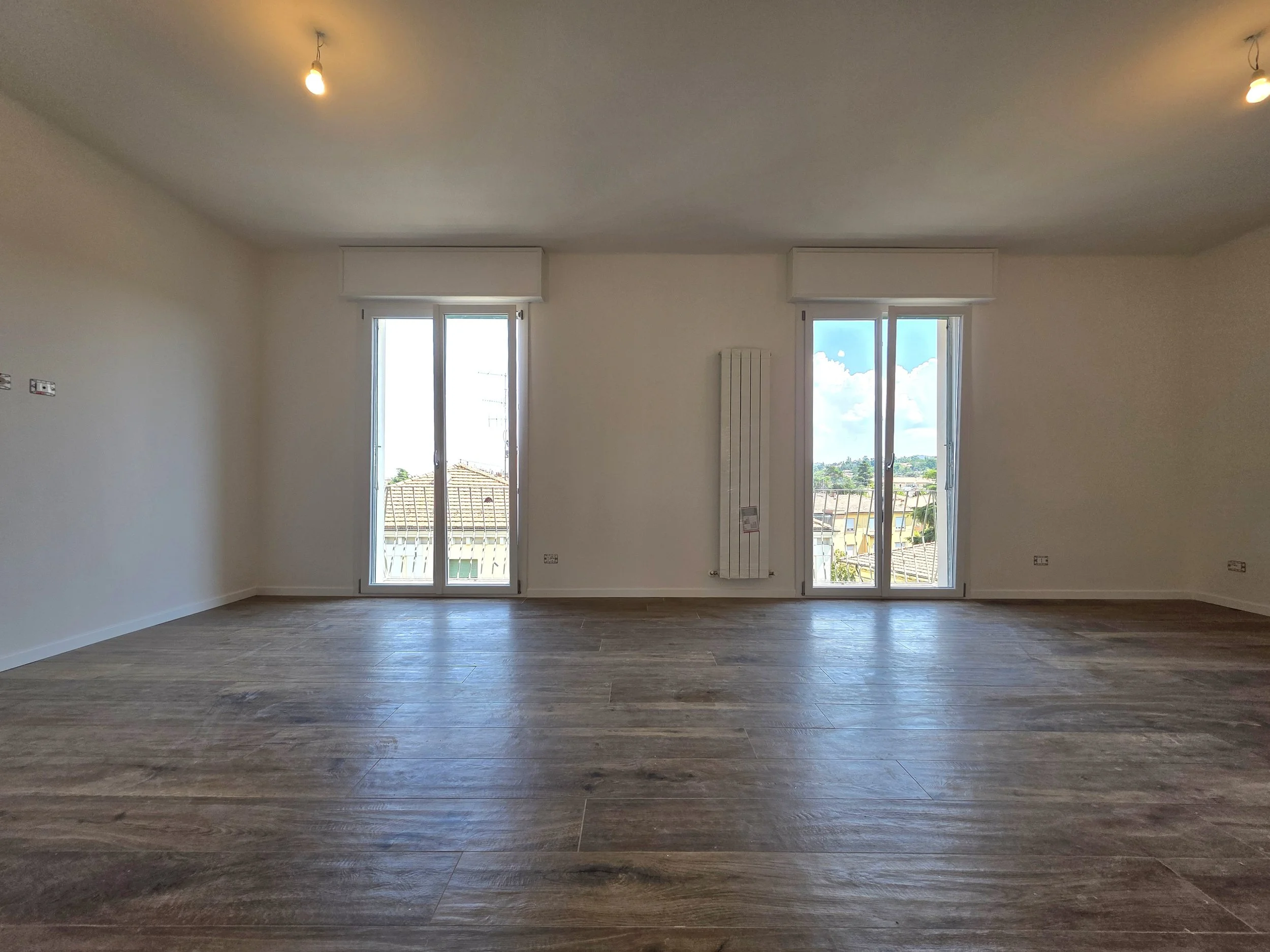 Soggiorno vuoto con pareti bianche, due finestre grandi con vista su case e cielo blu, pavimento in legno, soffitto con due luci spente.