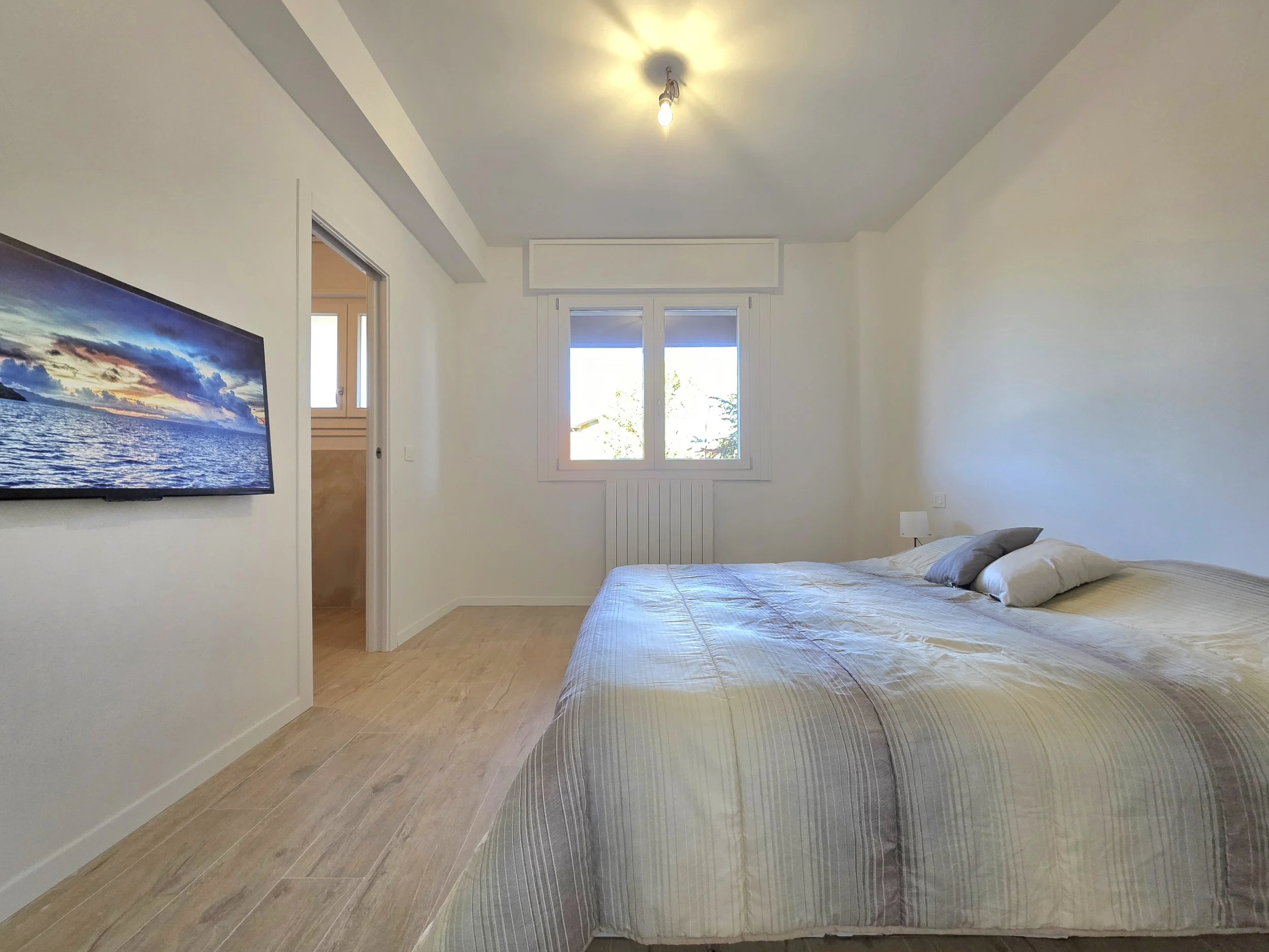 Camera da letto con letto matrimoniale, finestra che lascia entrare luce naturale, televisor appoggiato al muro, luce al soffitto, pavimento in legno chiaro.