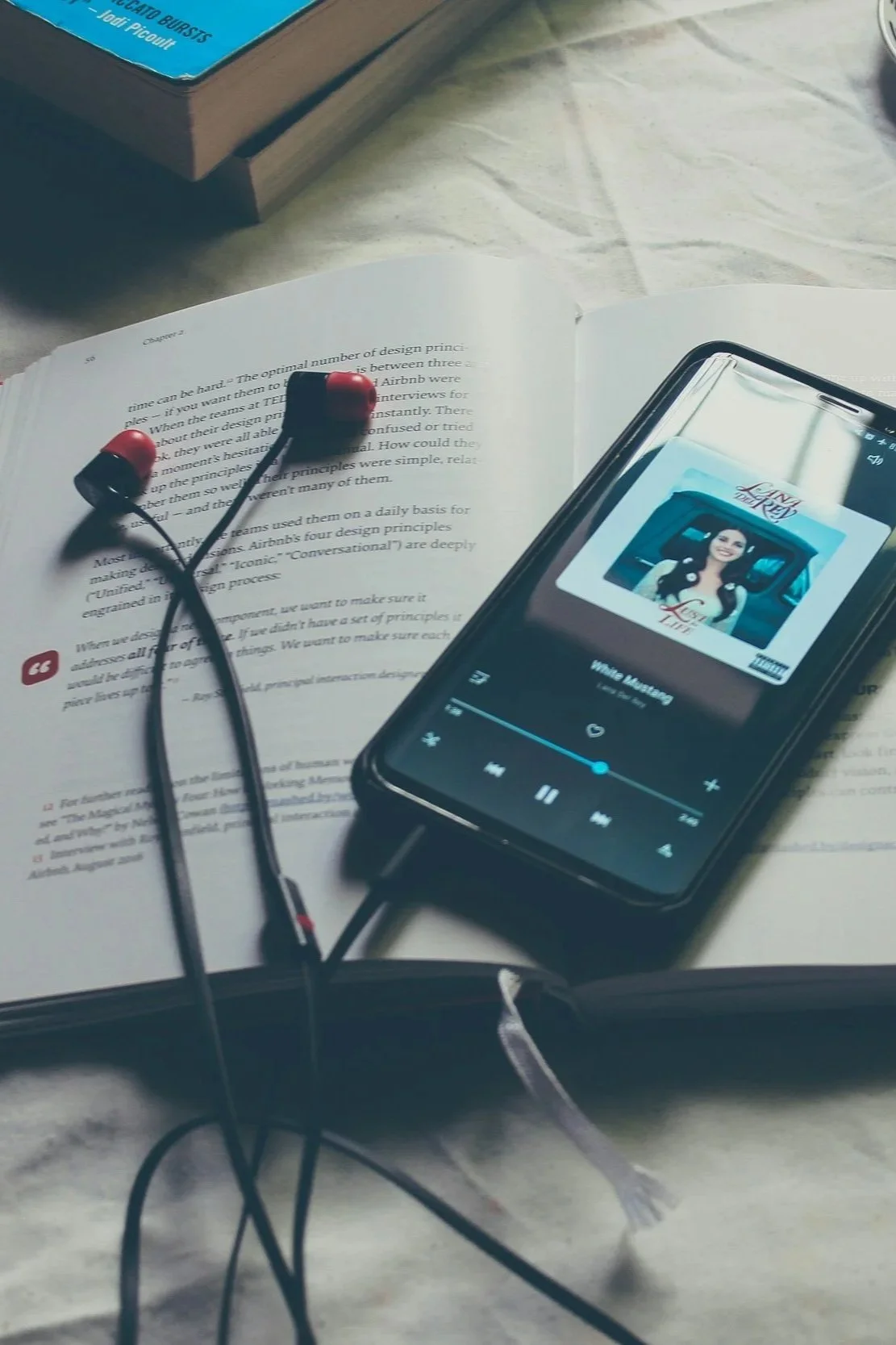 A smartphone playing music with album cover of a woman and text 'Yes I Like' on a bed with a book and red and black wired earphones resting on an open page of the book.