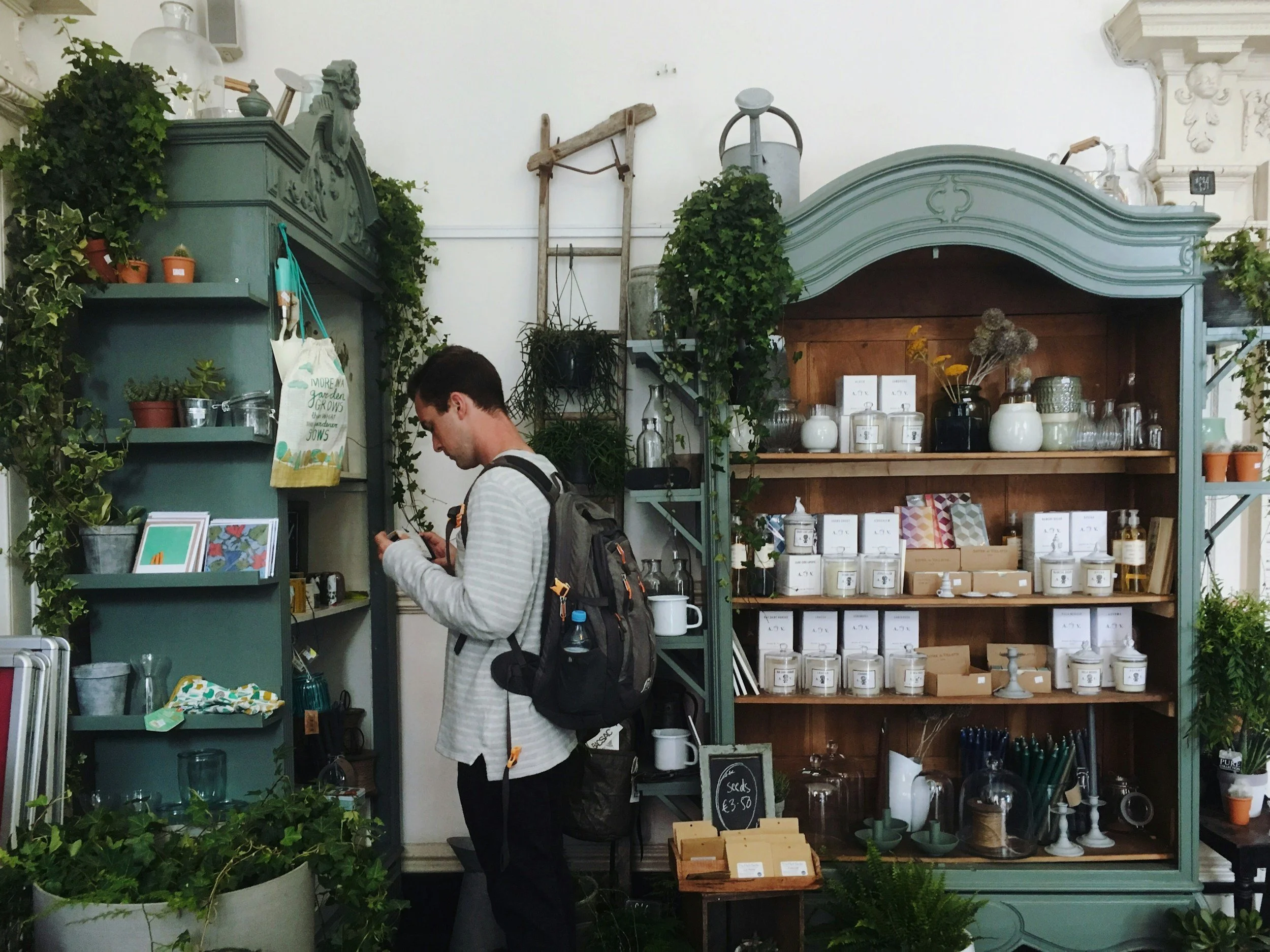 A man with dark hair and a backpack shopping in a gift shop store. The store has green and wooden shelves filled with bookish gifts, plants, jars, and decorative items.