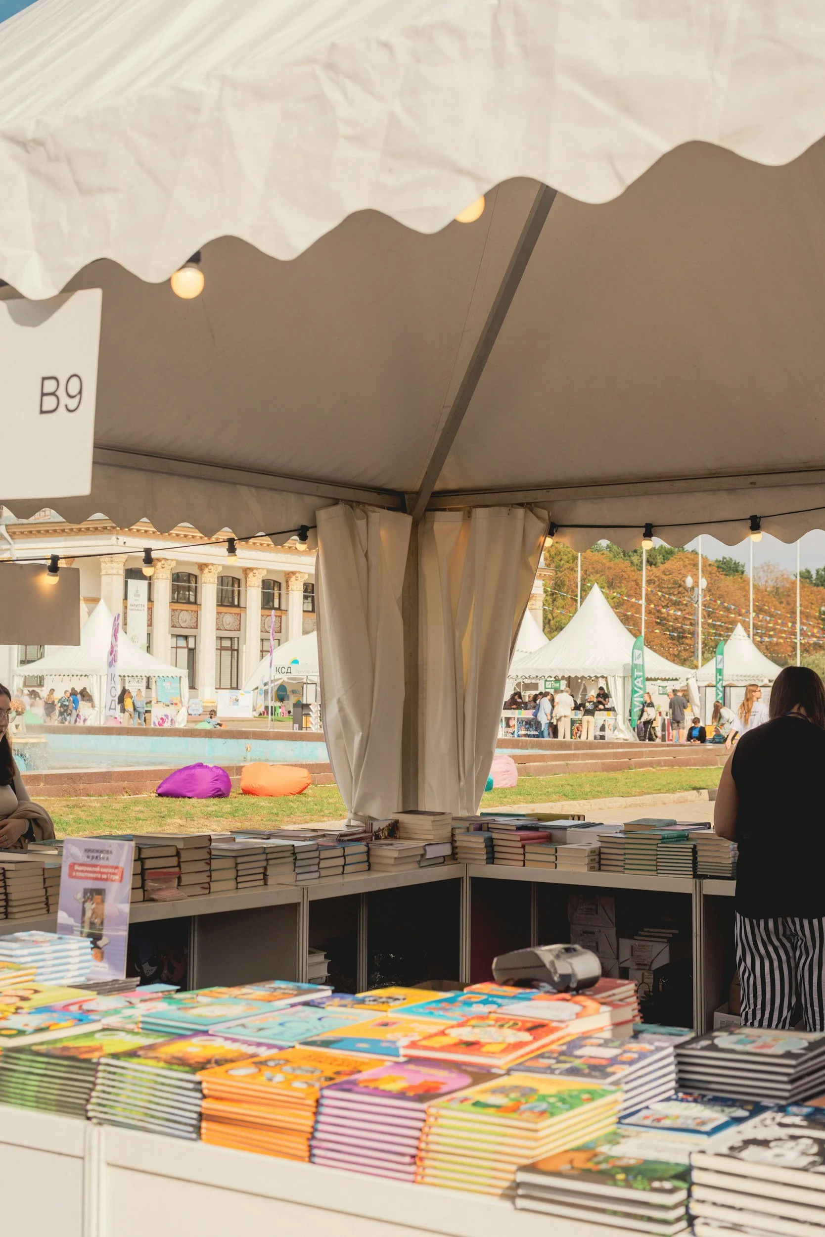A book stall at an outdoor event with stacks of colorful books on tables under white tents. In the background, there are historic buildings, other tents, and people walking around.
