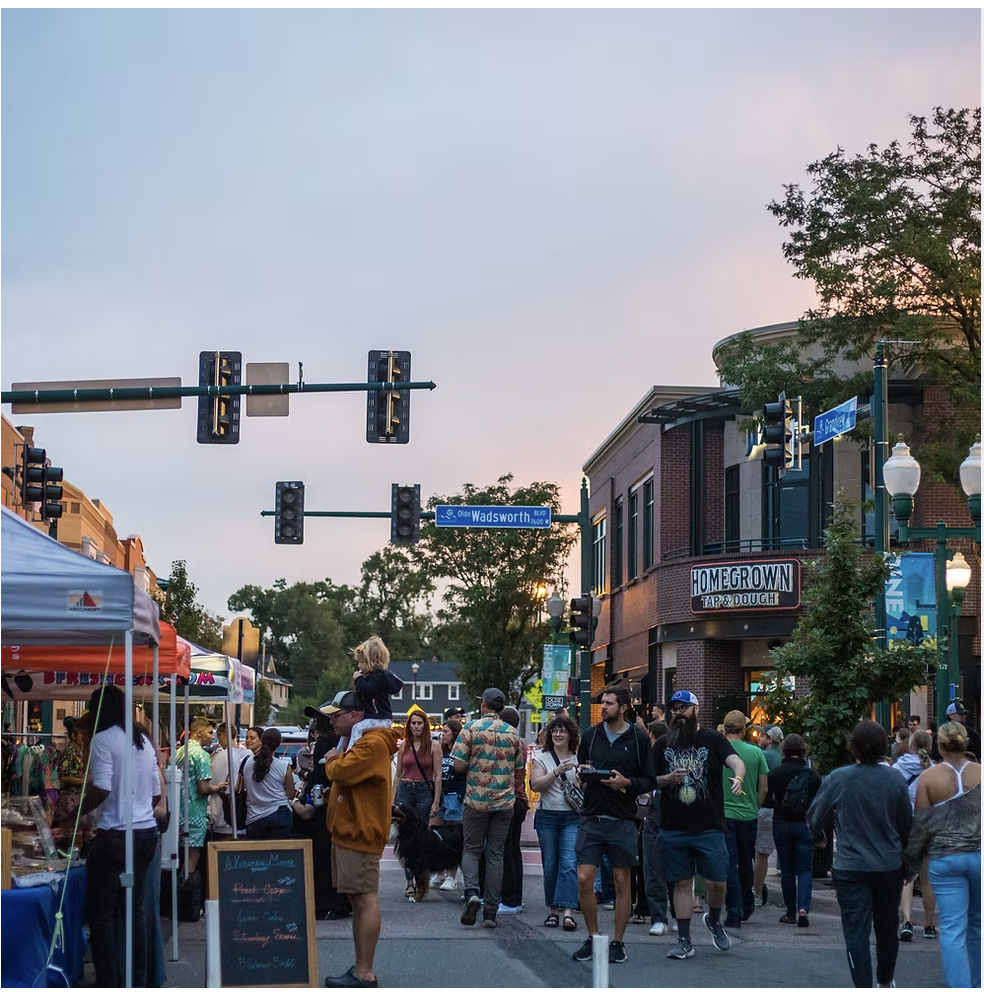 Crowd of people walking and shopping on the streets of Olde Town Arvada during the Second Saturday Concert Series outdoor event at dusk, with tents, street signs, and storefronts visible.