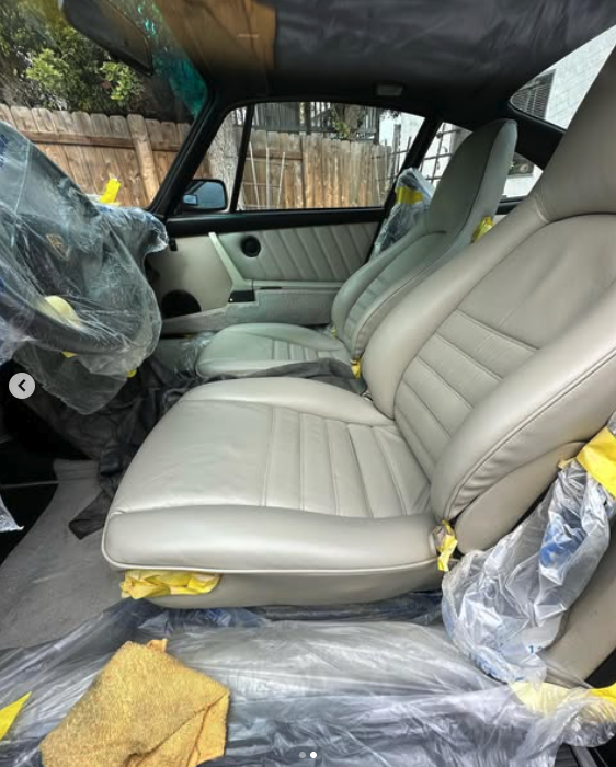 Interior of a car with beige leather seats and protective plastic covering, parked outdoors near a wooden fence.