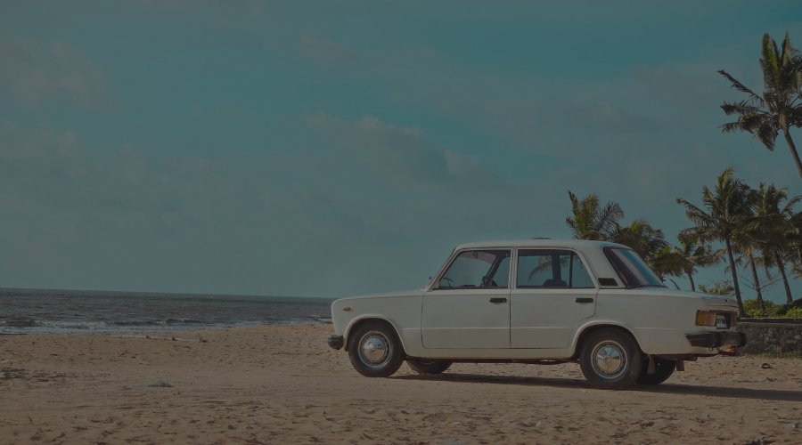 A vintage white car parked on a sandy beach with palm trees and the ocean in the background.
