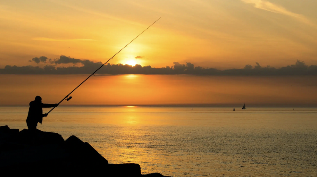 Silhouette of a person fishing with a long rod at sunrise over a calm sea, with distant sailboats and clouds in the sky.