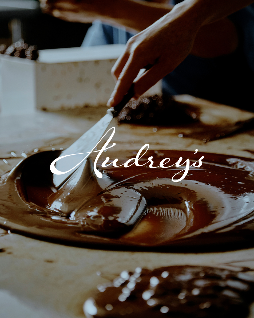 Close-up of a person spreading melted chocolate with a spatula on a wooden surface, with the text "Audrey's" overlayed.