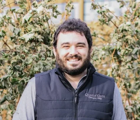 A smiling man with dark curly hair and a beard, wearing a black vest over a gray shirt, standing outdoors in front of a background of plants or vines.