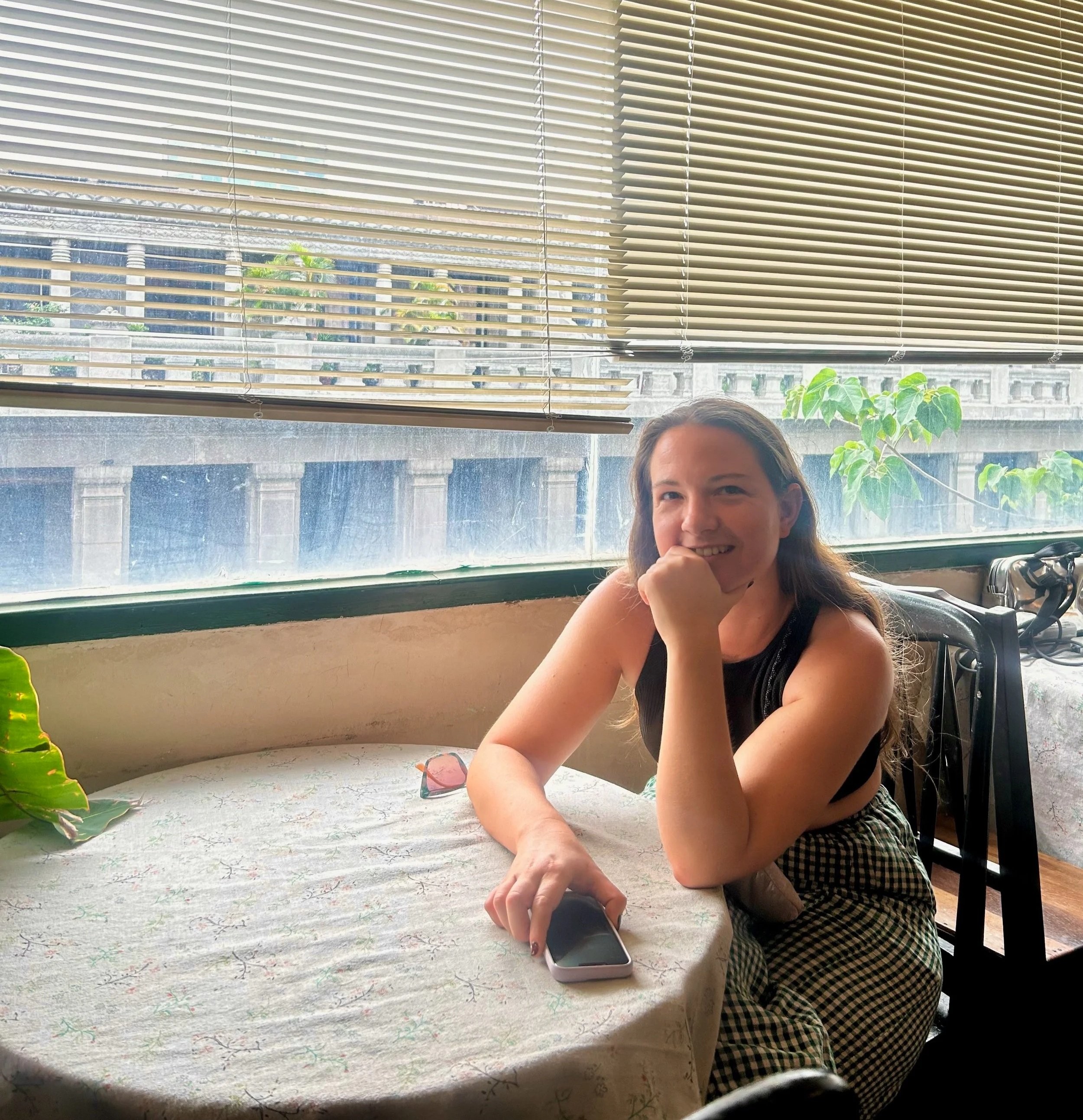 A young woman with long brown hair smiling and resting her chin on her hand while sitting at a table in a cafe. She is holding a smartphone in her right hand and wearing a black sleeveless top and checkered pants. The table has a light-colored tablecloth, and there are plants on the windowsill behind her with closed blinds and a view of a building exterior outside.