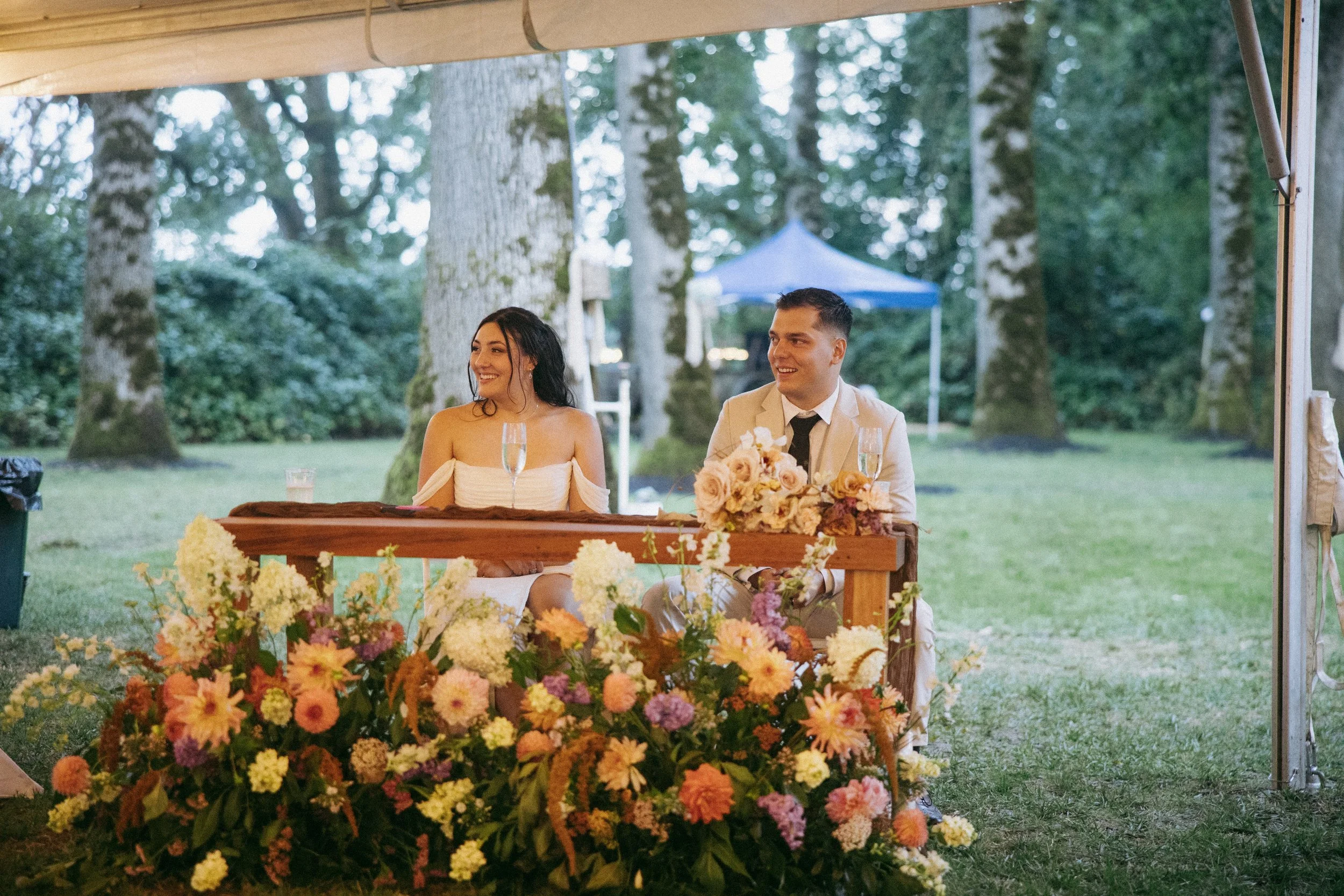 A couple sitting at a decorated table outdoors, smiling, with a garden and trees in the background during a wedding celebration.