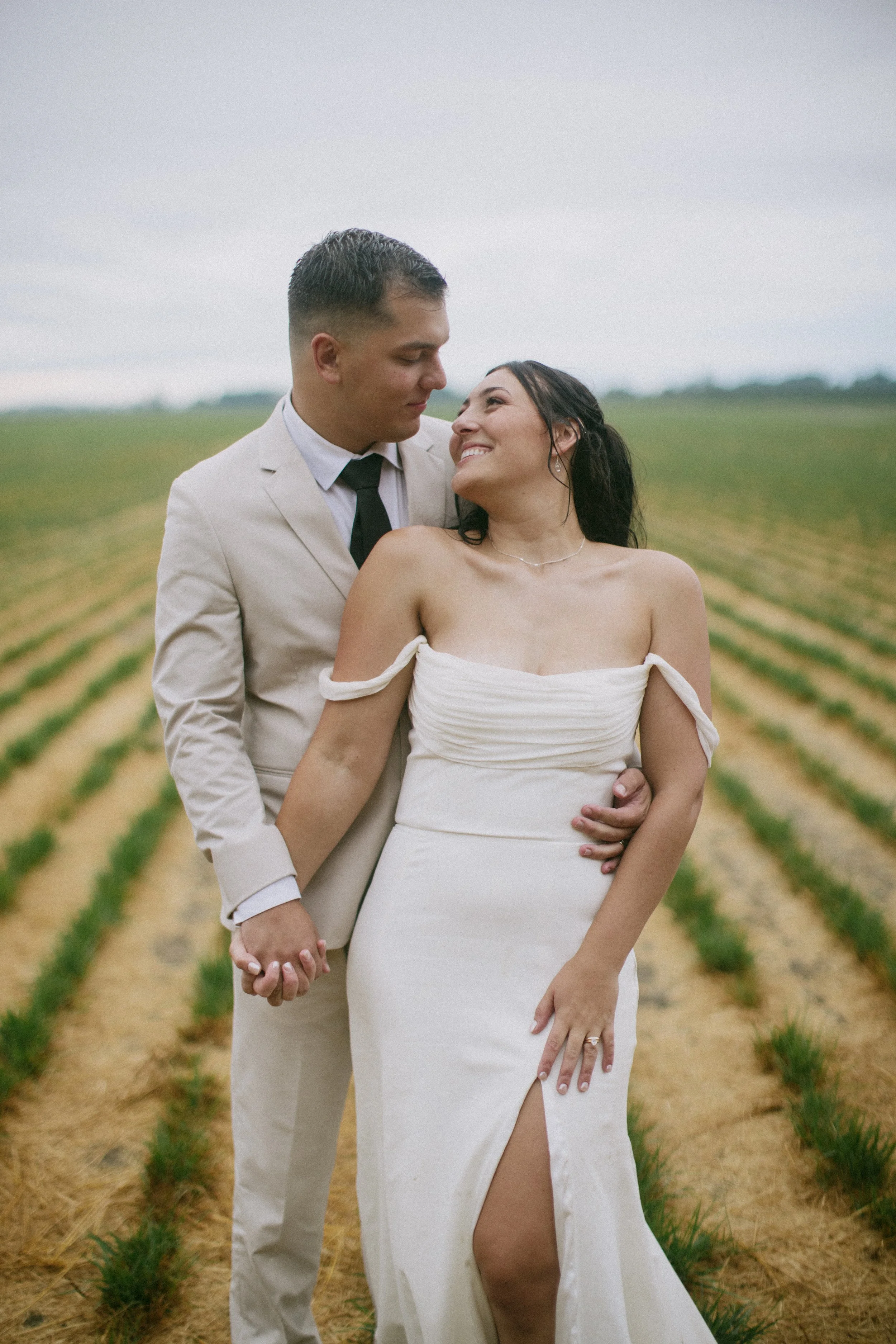 A couple in wedding attire standing in a field, holding hands, looking at each other, with a cloudy sky in the background.