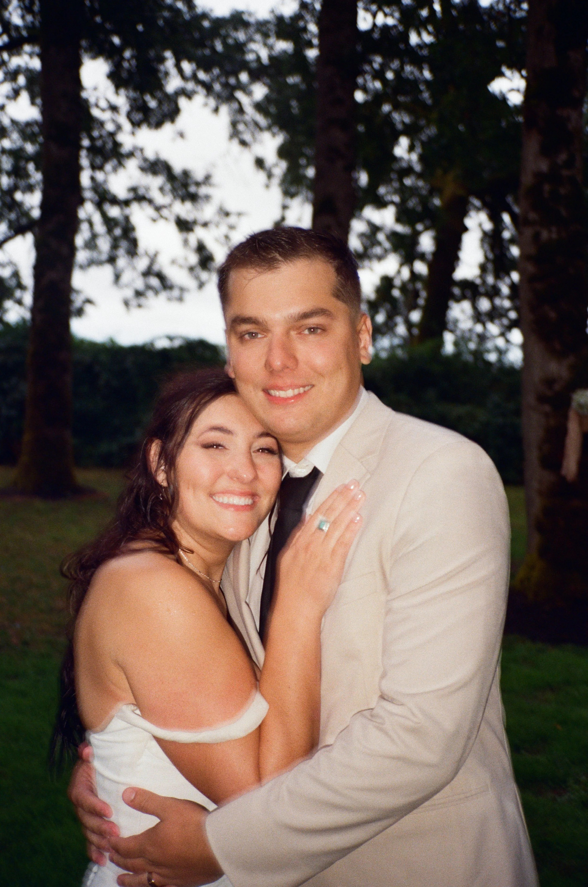 A couple embraces outdoors, smiling, with trees in the background during evening.