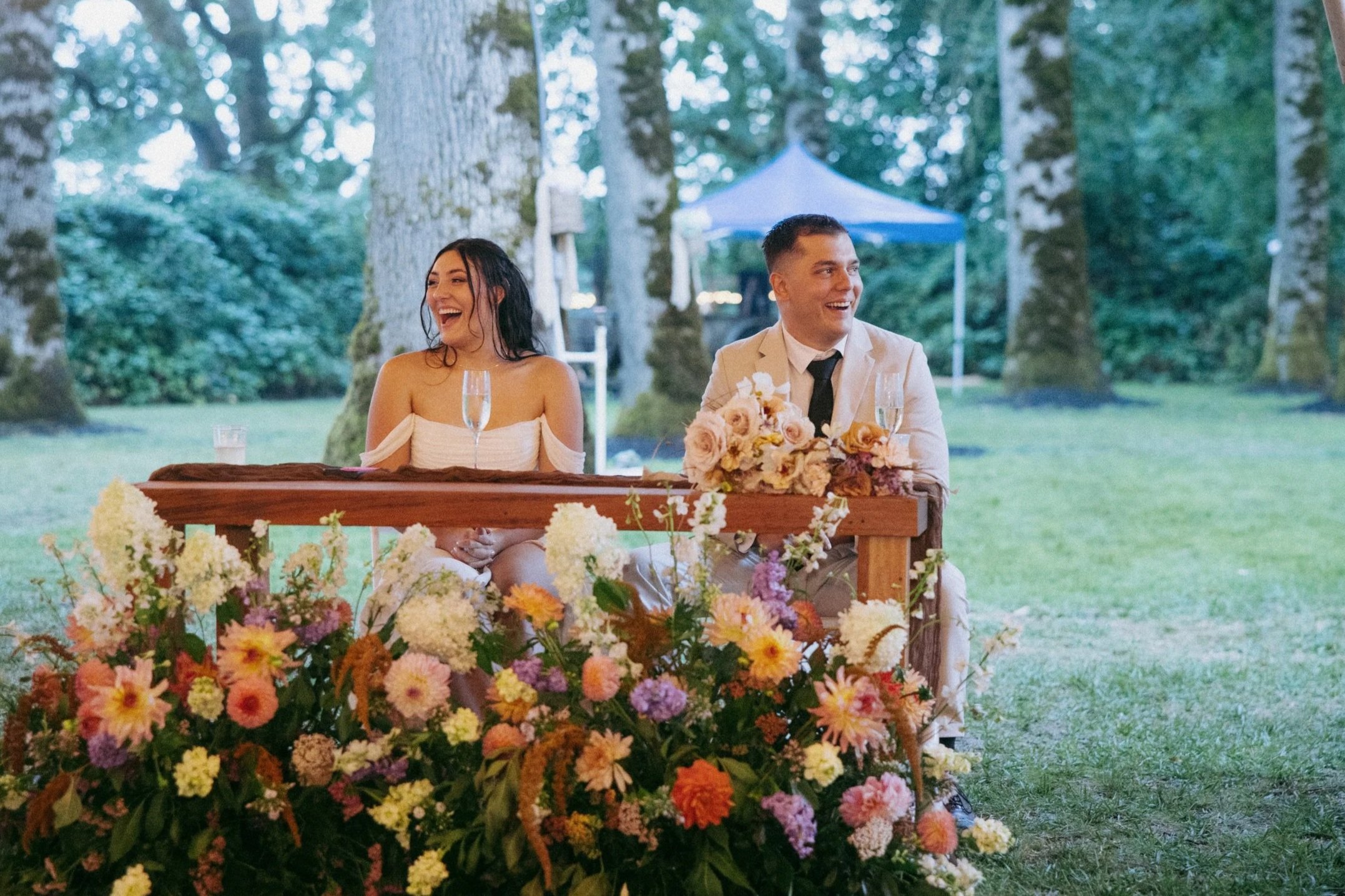 A joyous couple sitting at a decorated table outdoors during a celebration, with a backdrop of a forest and a blue canopy, surrounded by colorful flowers.