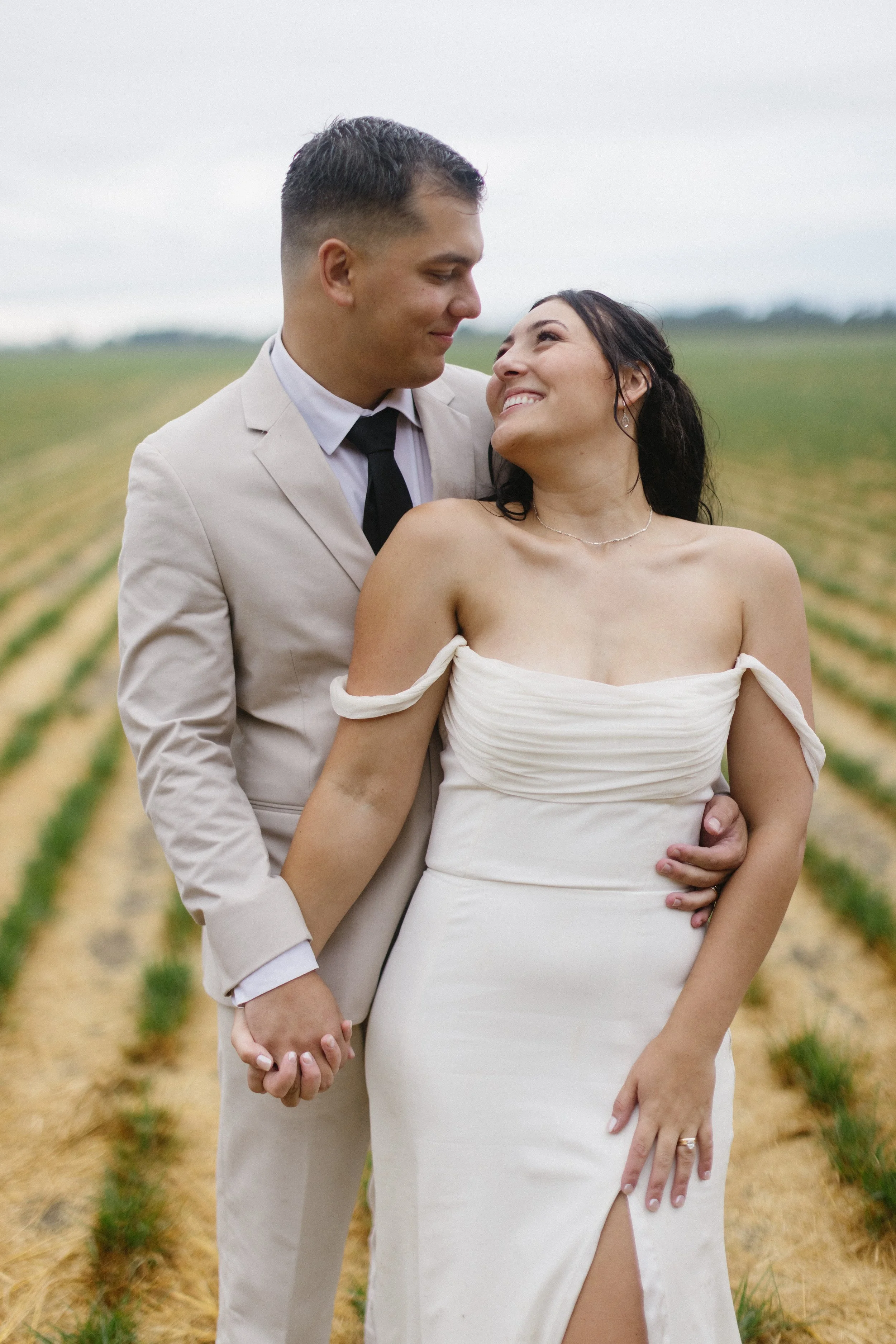 A couple in wedding attire holding hands and smiling at each other outdoors on a cloudy day in a field of small plants.