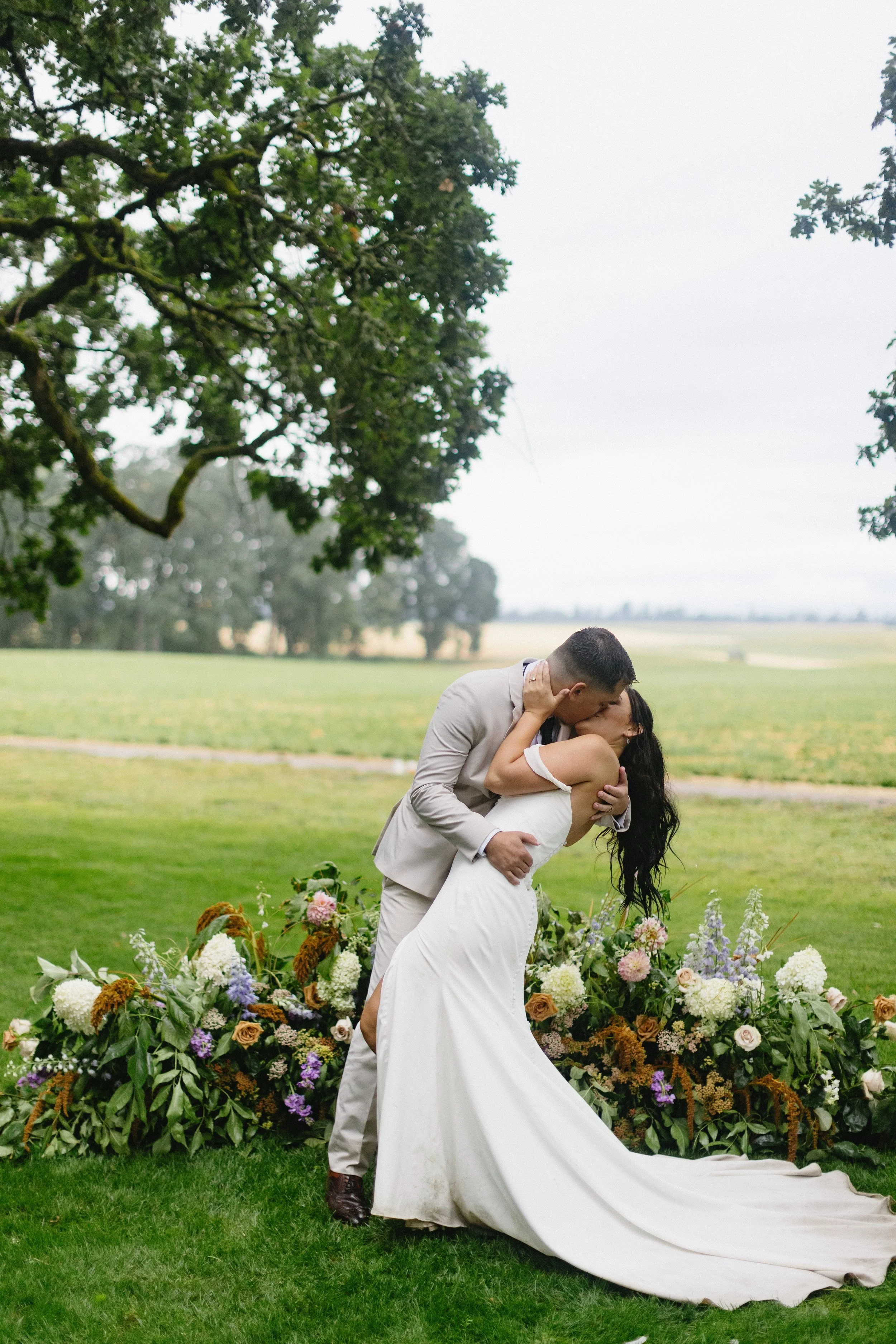 A wedding couple kissing outdoors on a grassy field with a large flower arrangement and trees in the background.