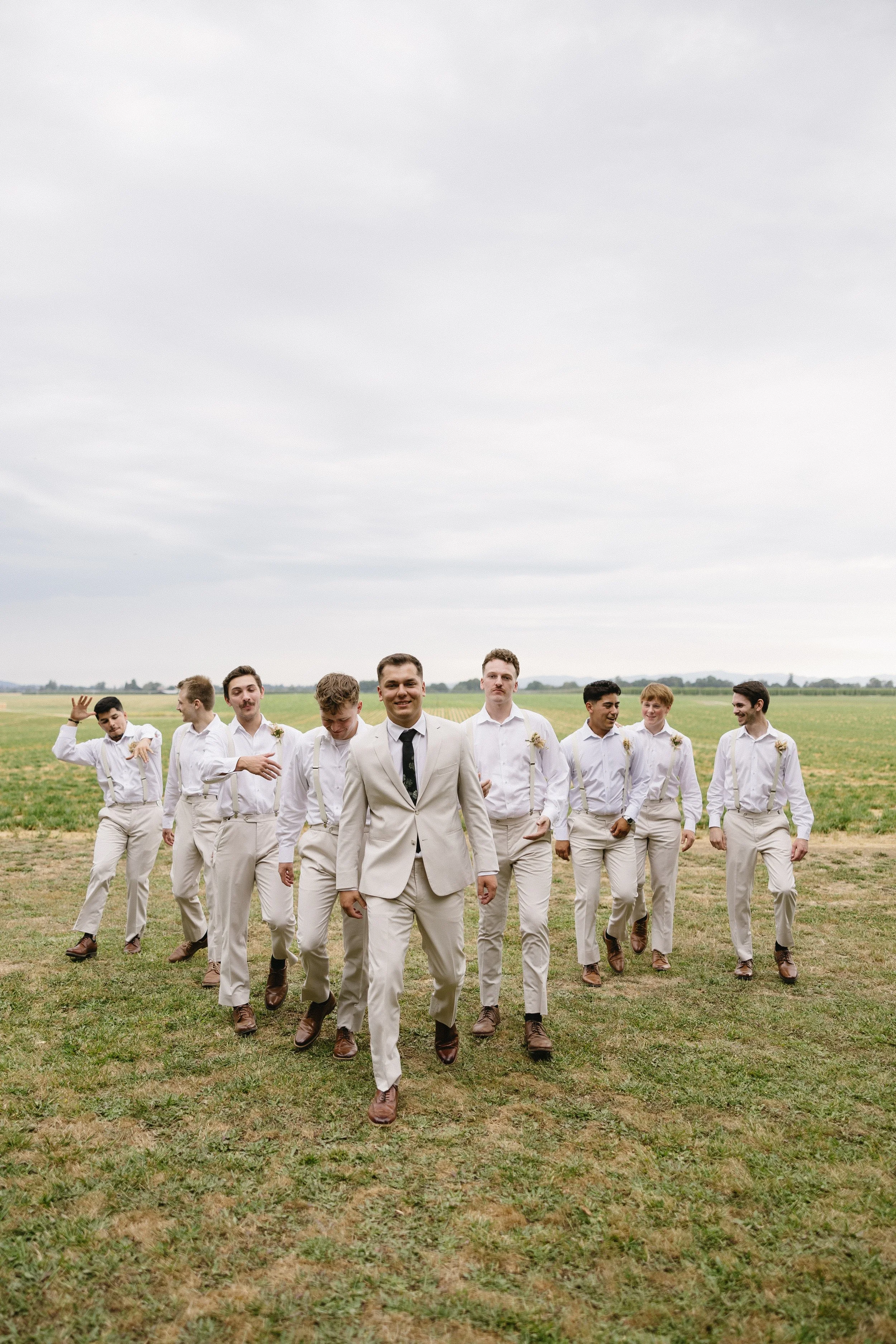 Group of men in cream-colored suits walking across a grassy field on an overcast day, with some waving and smiling.