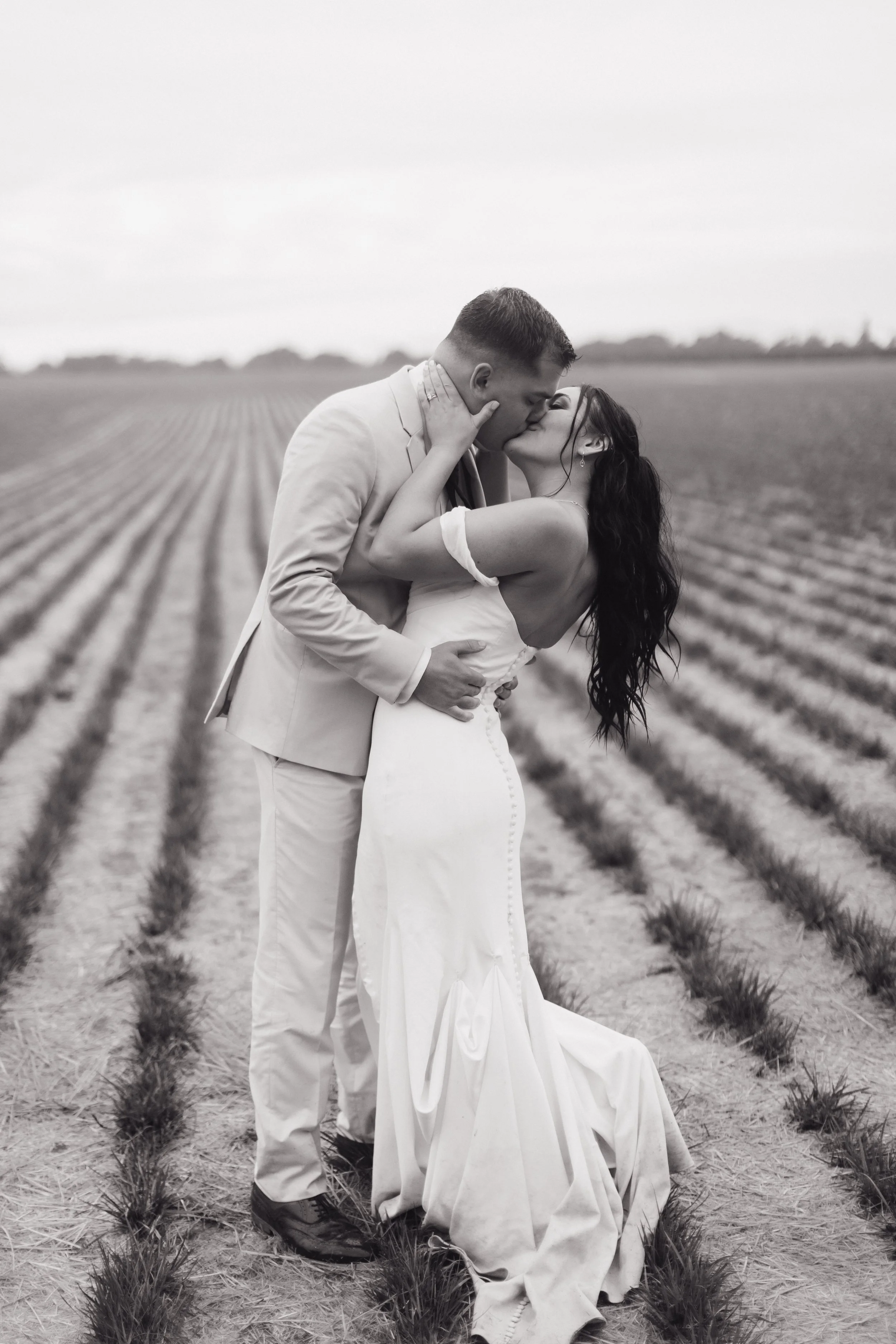 A black and white photo of a couple in wedding attire sharing a kiss in a field with rows of plants.