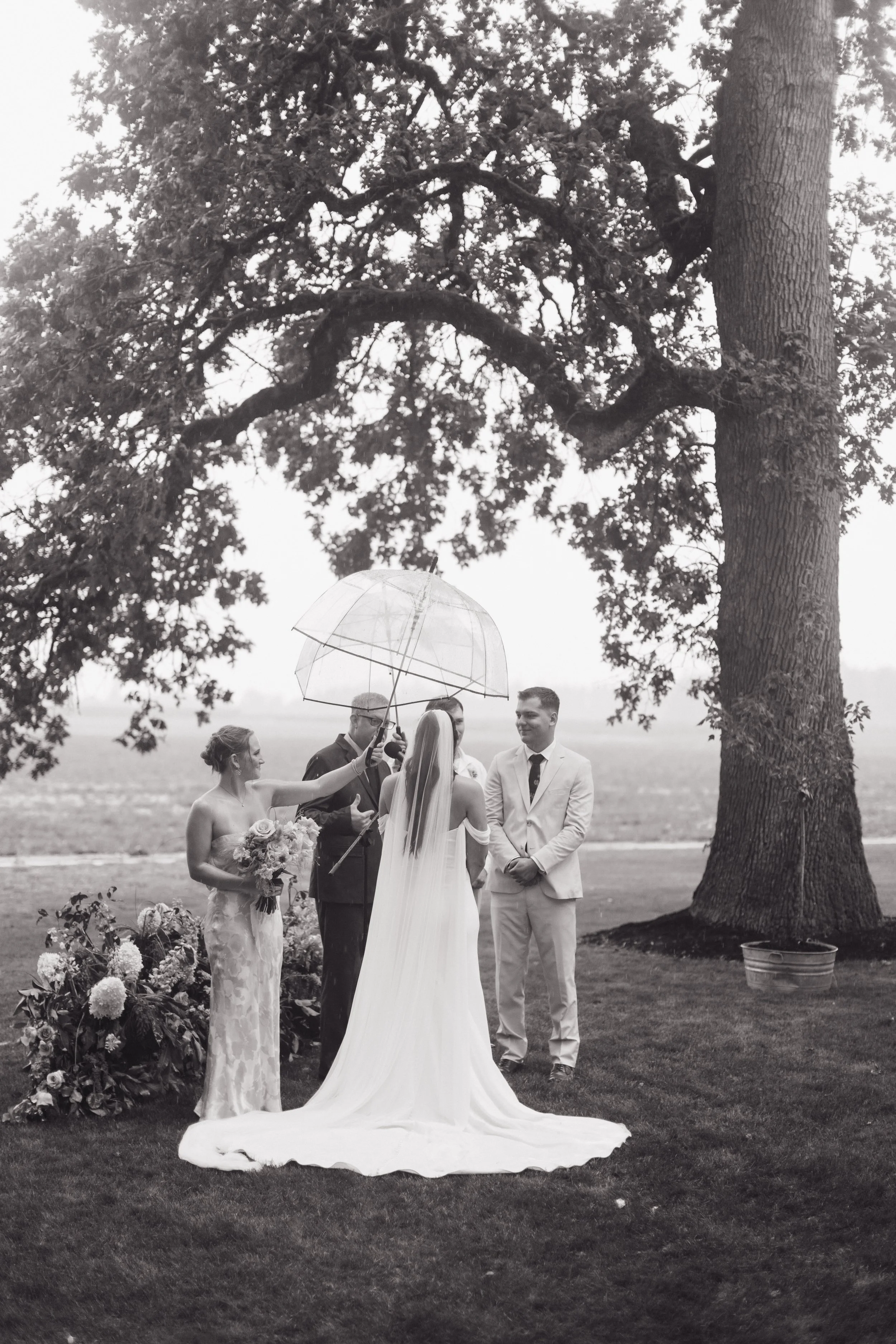 A wedding ceremony taking place outdoors with the bride and groom standing under a large tree. The bride is wearing a long wedding dress and veil, holding a bouquet, while the groom is in a light-colored suit. An officiant is holding an umbrella over
