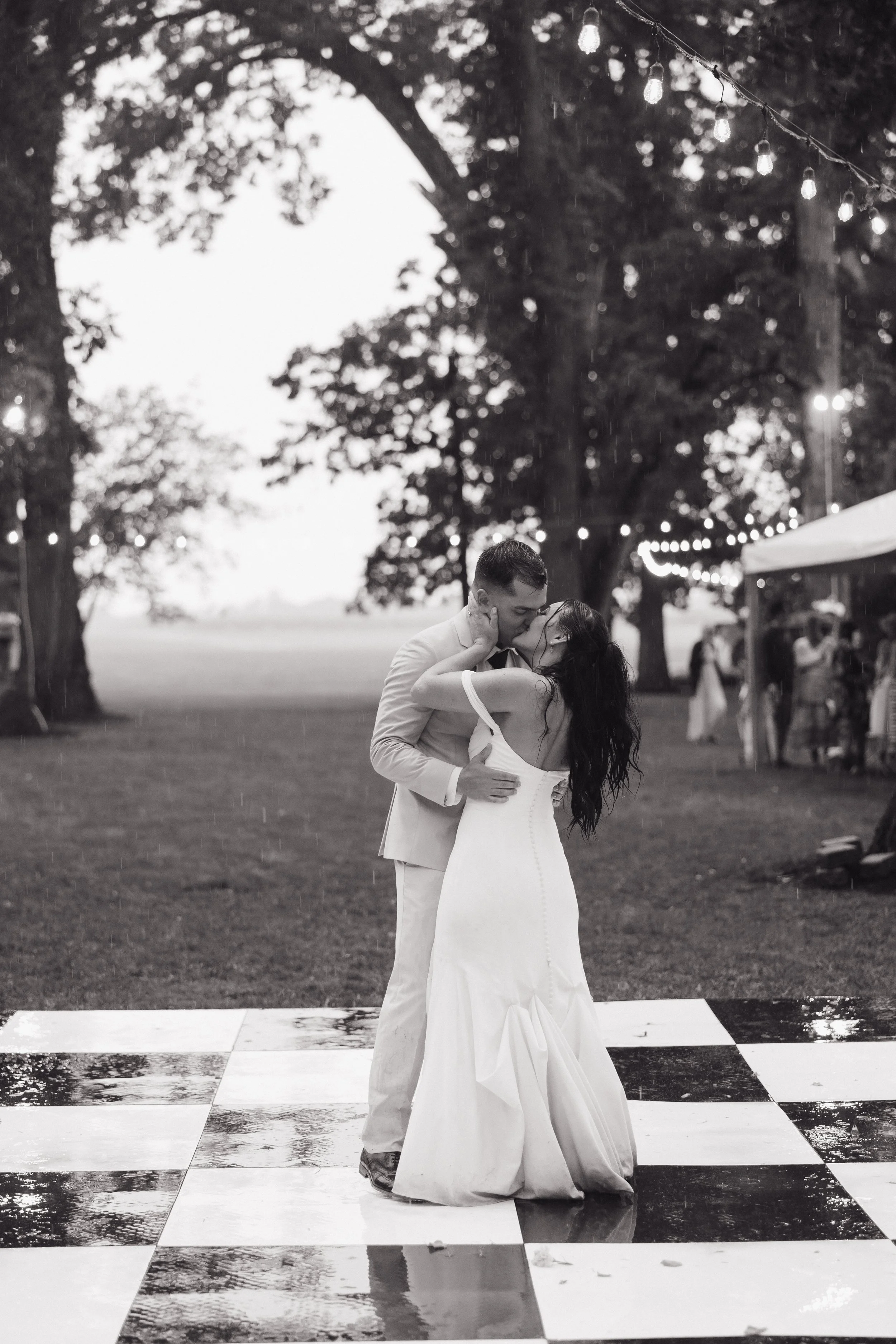 A couple dancing outdoors on a black-and-white checkered dance floor during a rainy wedding reception, with string lights overhead and trees in the background.