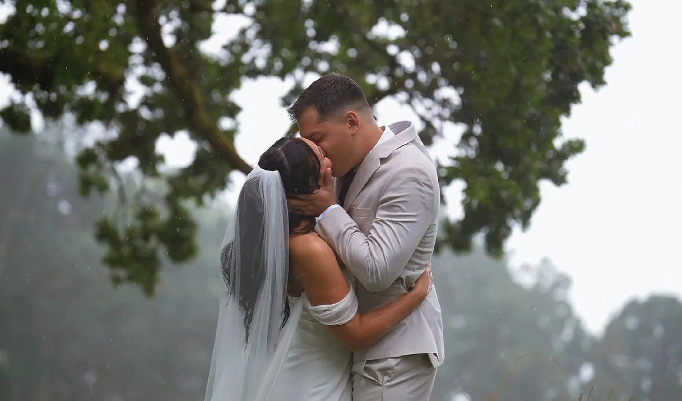 A bride and groom embrace and kiss outdoors in the rain, with trees in the background.