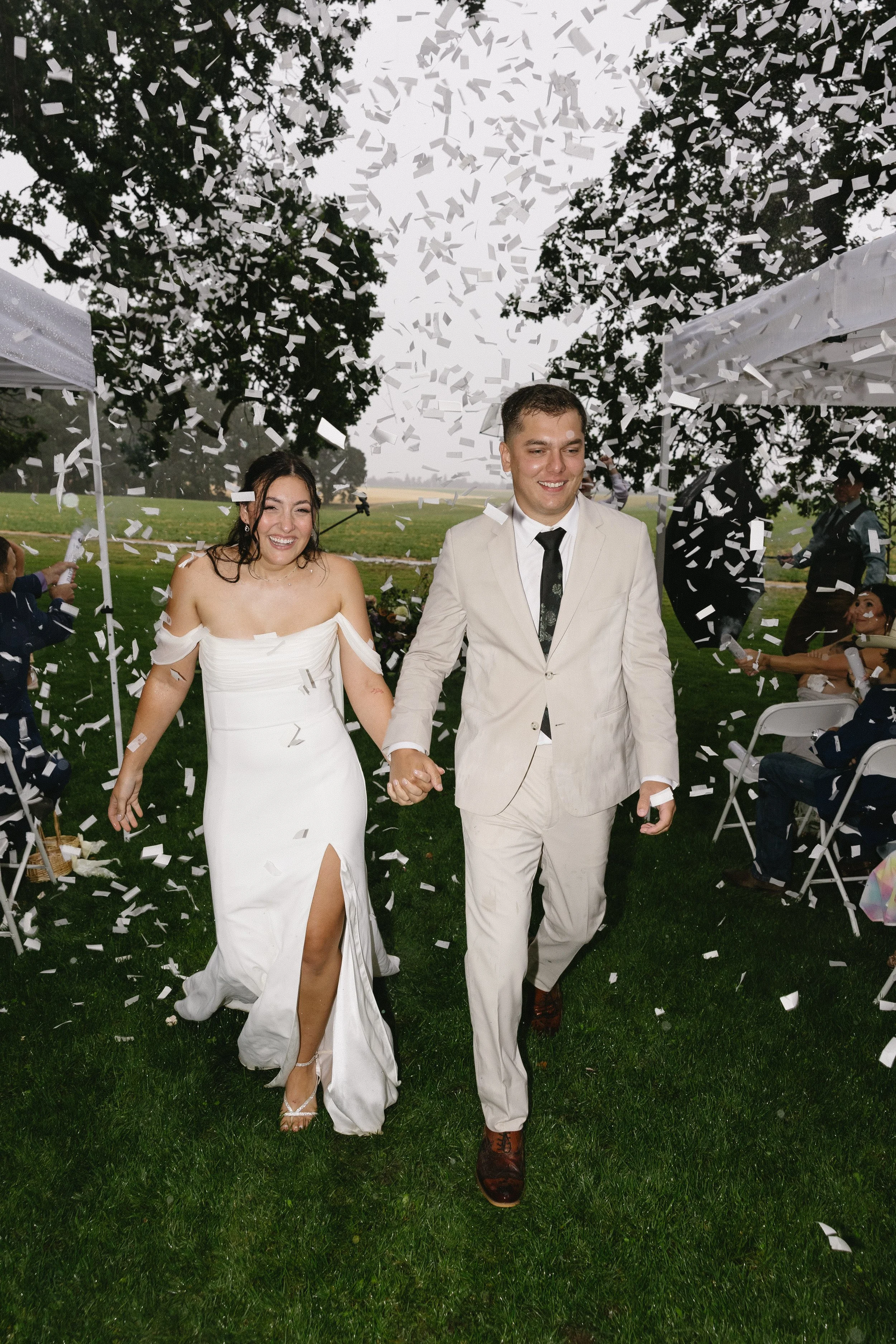 Bride and groom holding hands and smiling as they walk through falling confetti outdoors, surrounded by wedding guests under tents.