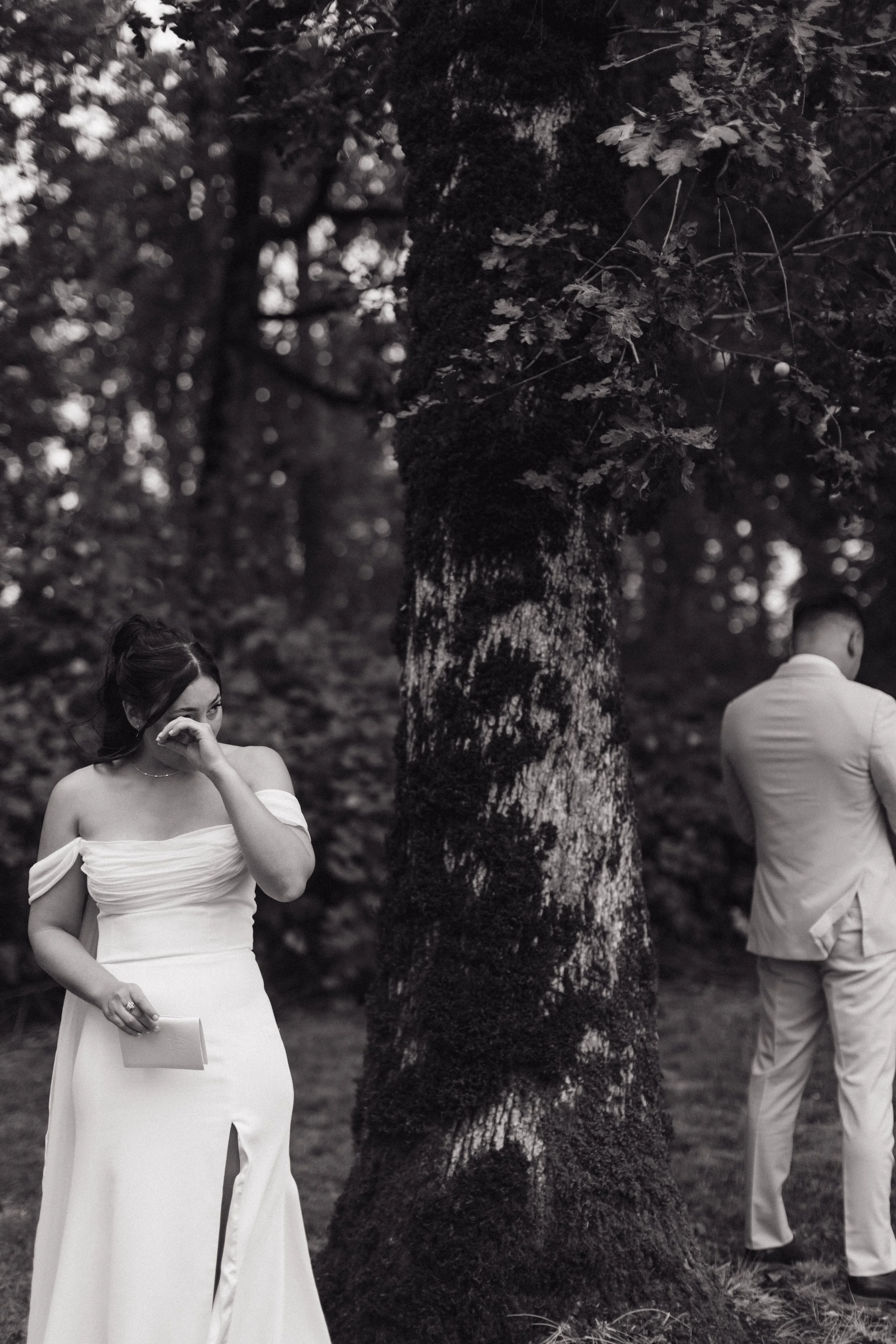 A black and white photo of a woman in a white strapless dress standing outdoors near a large tree, covering her face with her hand, with a man in a white suit standing nearby facing away.