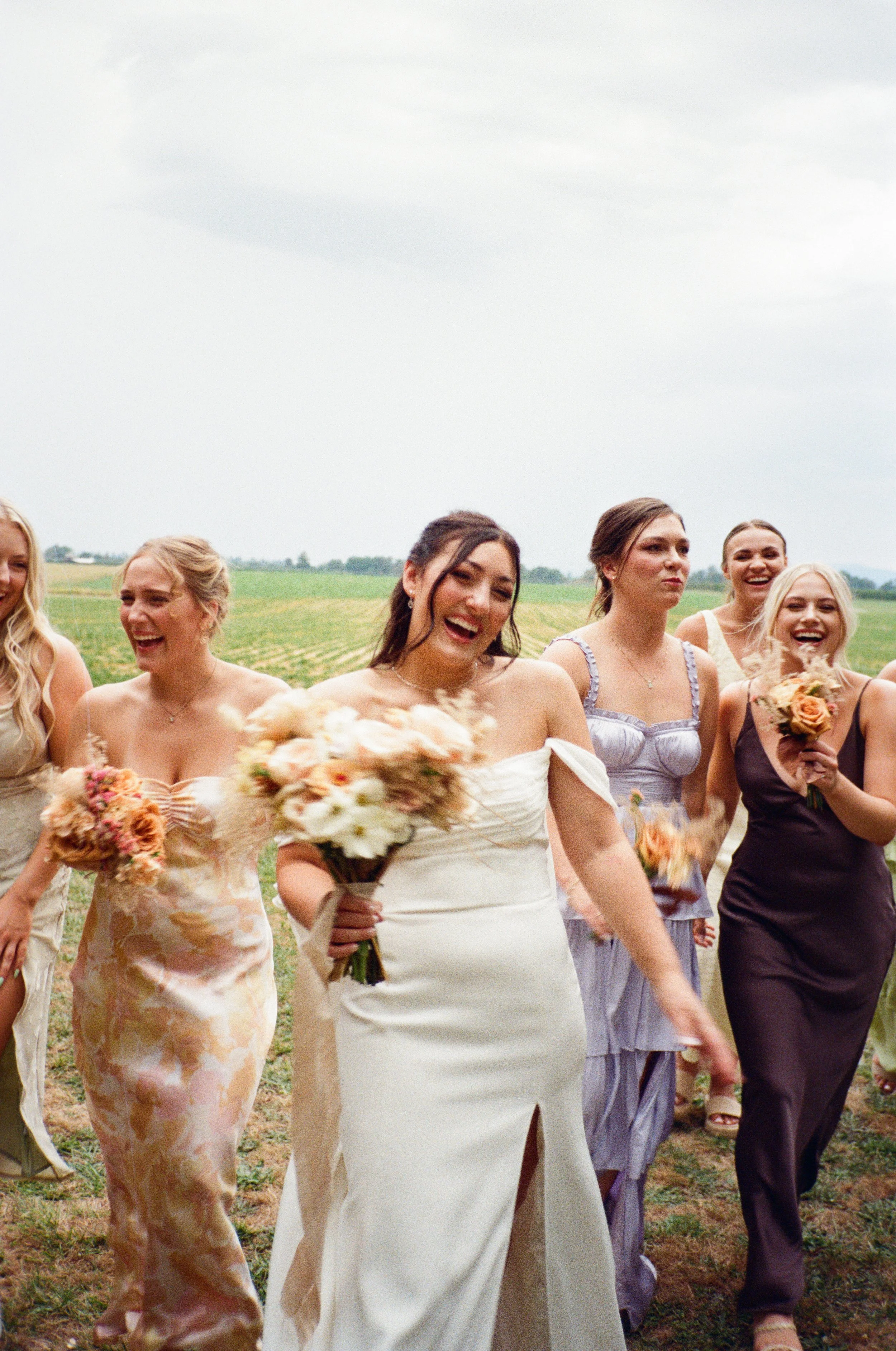 Group of women in dresses walking outdoors, holding bouquets, on a grassy field under cloudy sky.