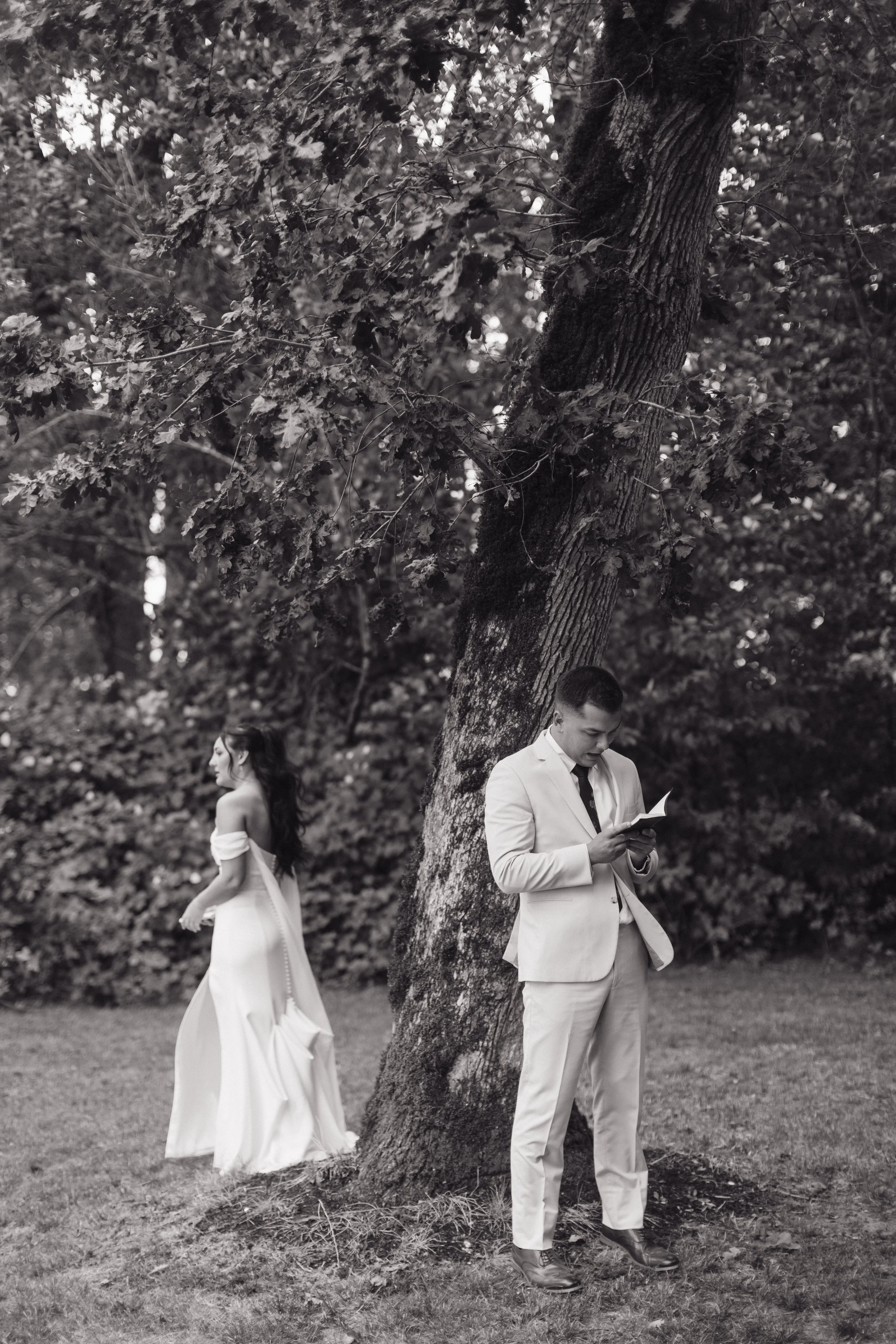 A black and white photo of a man in a suit reading a book or card under a large tree with a woman walking away in a flowing dress in the background.
