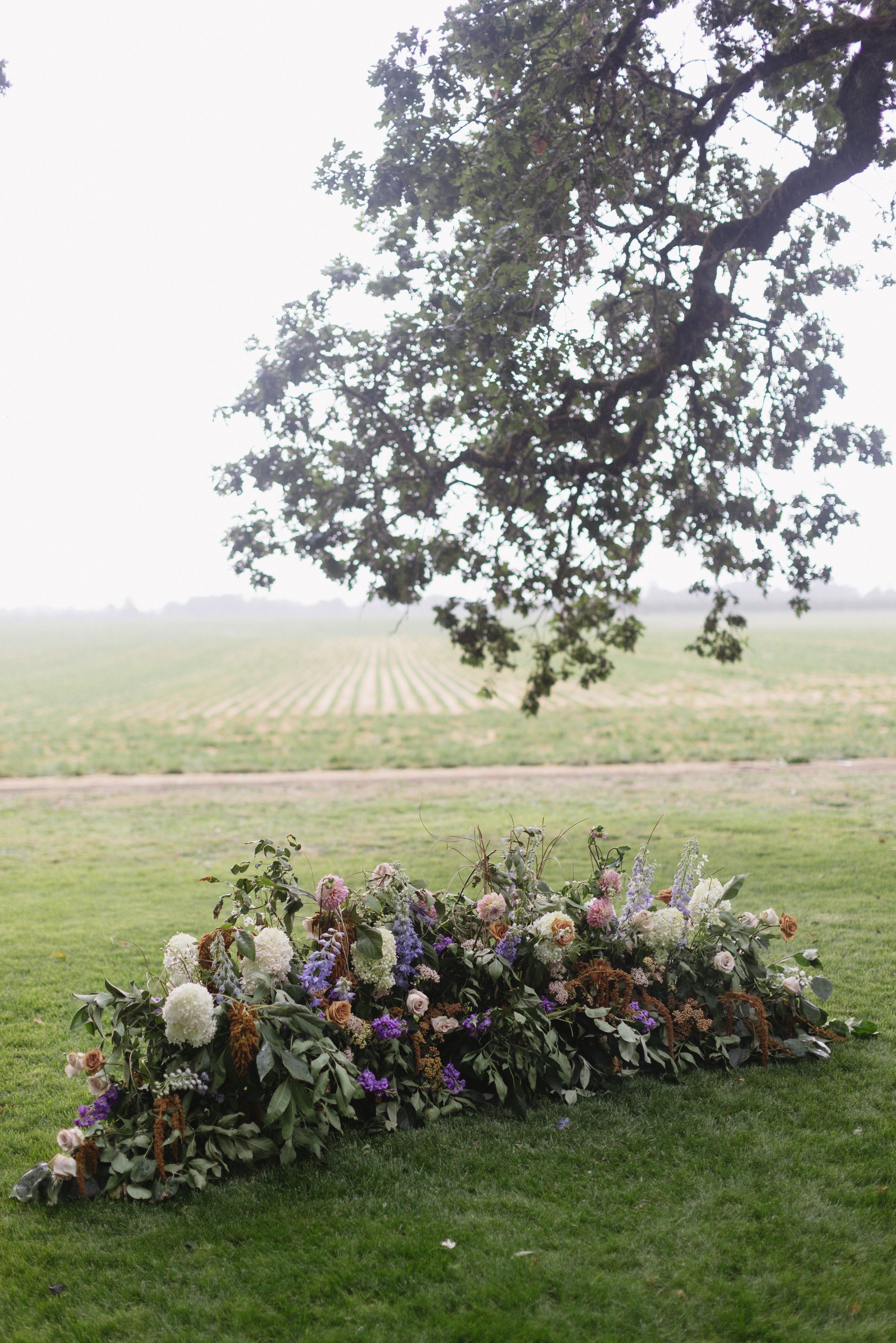 Flower arrangement with a mix of pink, purple, white, and brown flowers and greenery on a green lawn, with a tree overhead and a field in the background.