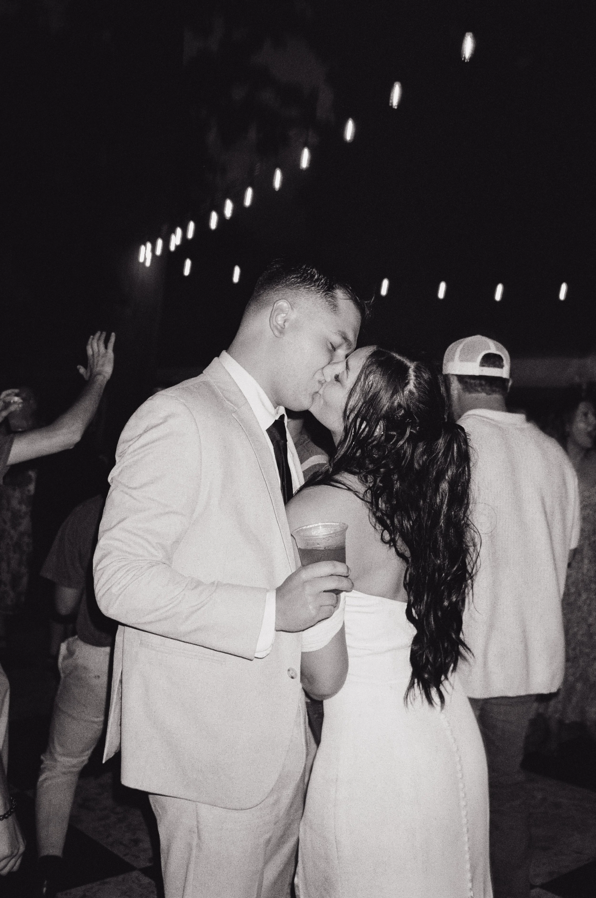A couple sharing a kiss at a wedding reception, with the man in a light-colored suit and the woman in a white dress, holding a drink.