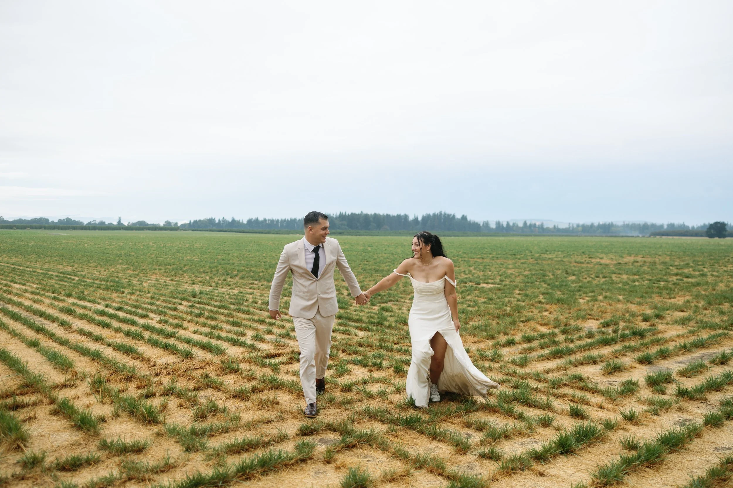 A couple dressed in wedding attire walking hand in hand across a green field.