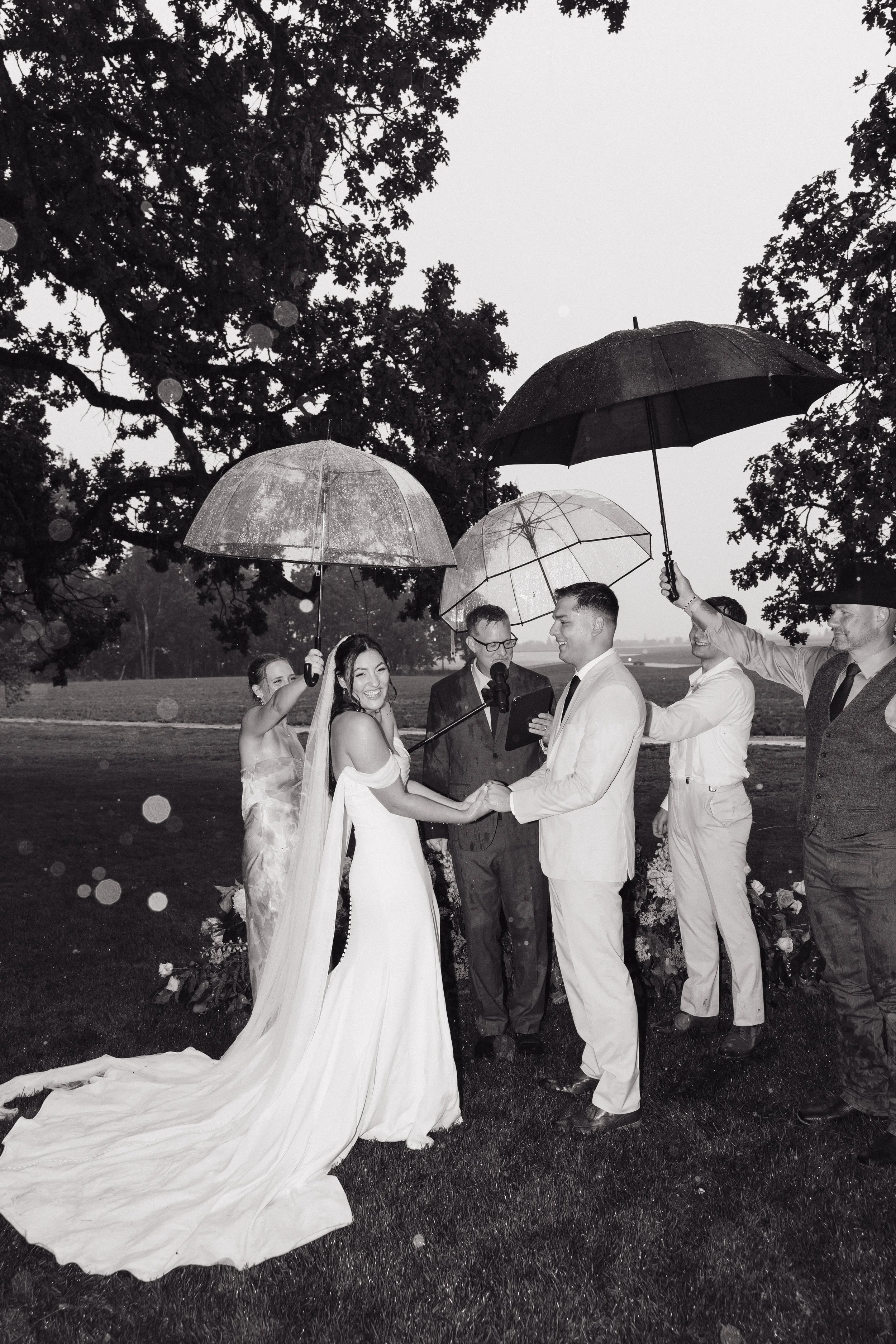 A wedding ceremony outdoors in the rain, with the bride and groom holding hands and smiling, surrounded by officiant and friends holding umbrellas.