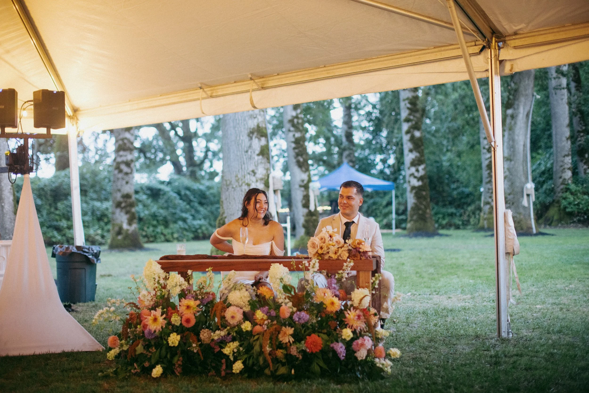 A couple sitting at a decorated table under a tent outdoors, surrounded by trees and greenery, with flowers on the table and a backdrop of a tent in the background.