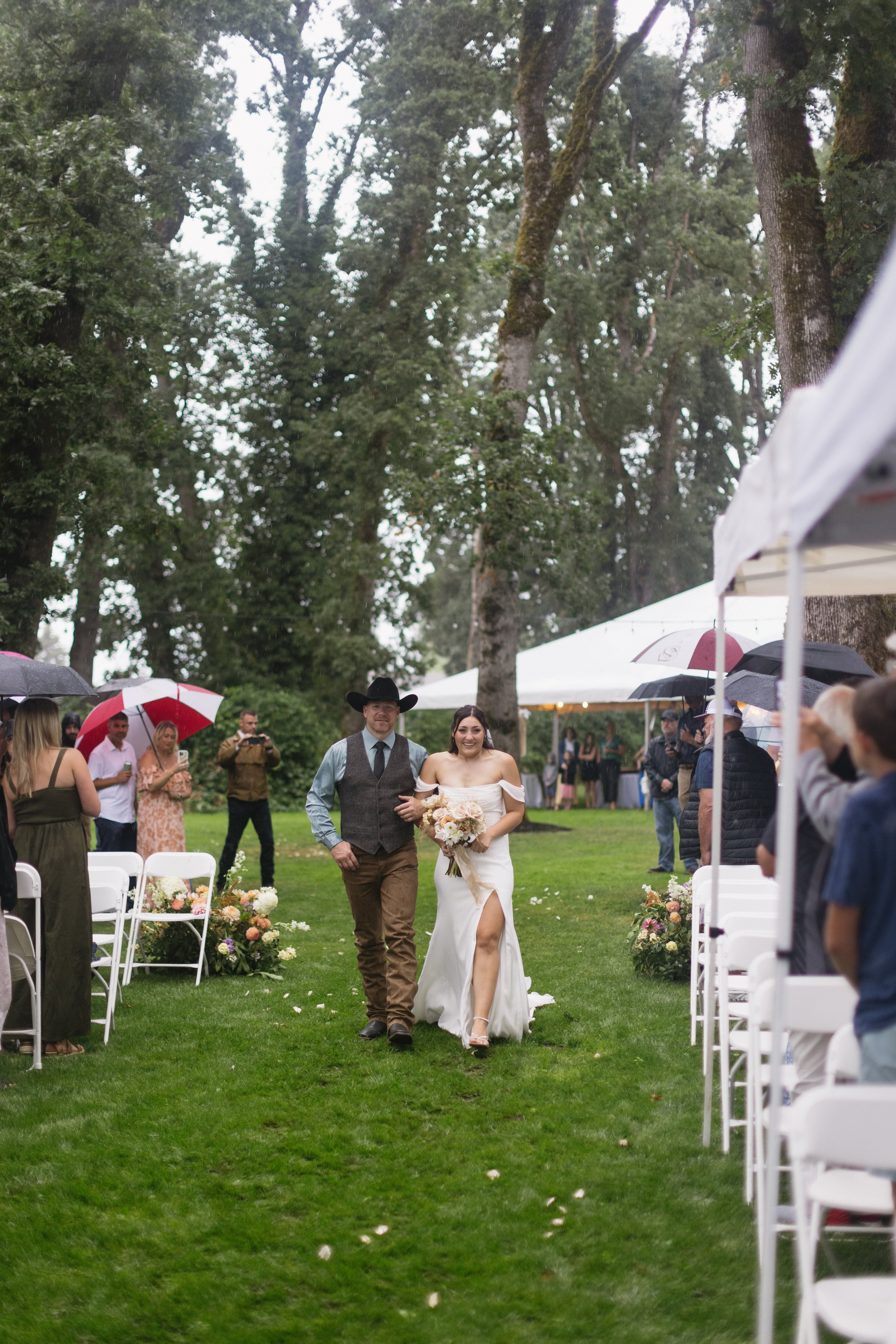 Bride walking down the aisle with her father at an outdoor wedding ceremony in a lush green forest, surrounded by guests holding umbrellas, in rainy weather.
