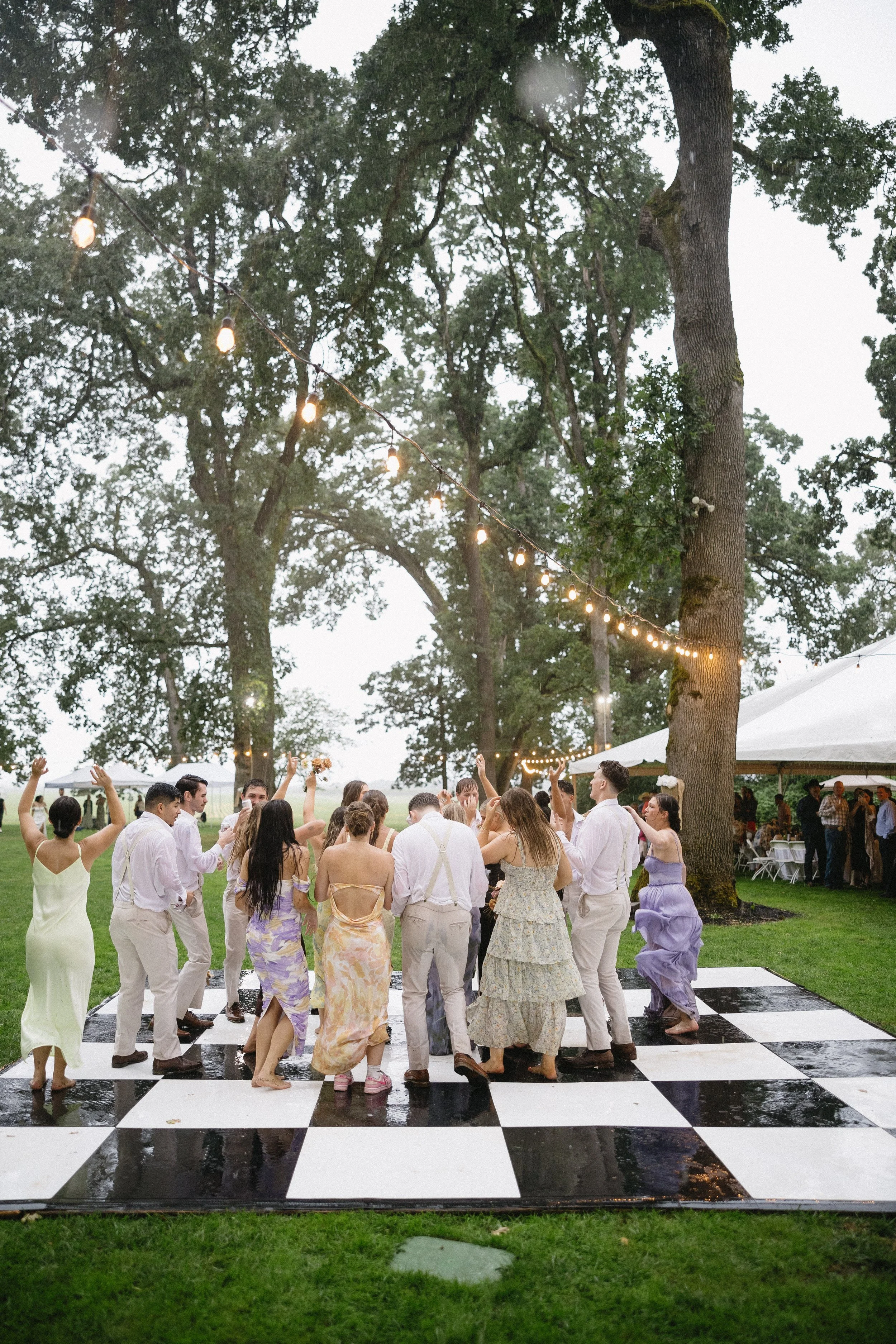 People dancing on a black and white checkered dance floor outdoors under string lights, with trees and a white tent in the background, during a celebration or party.
