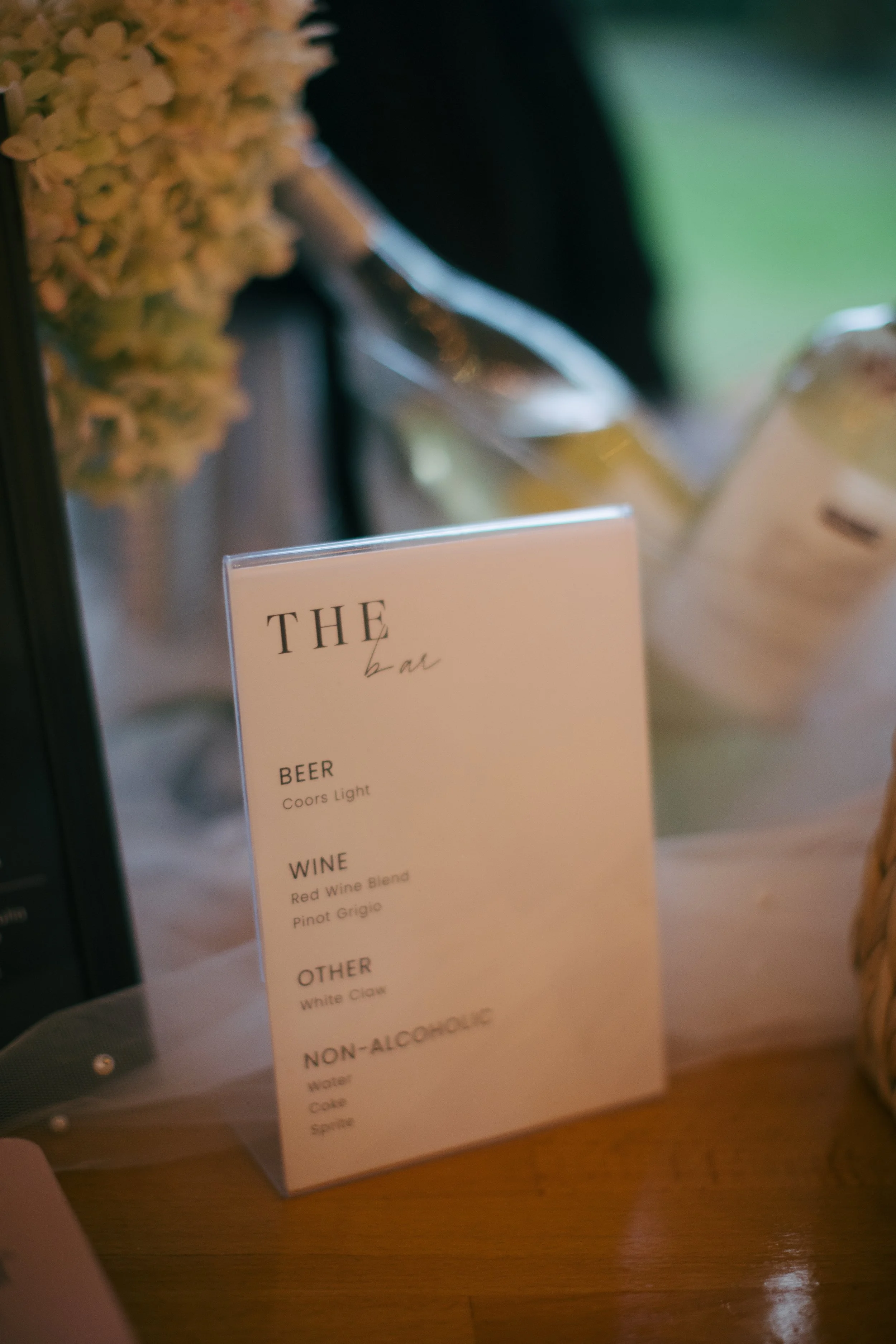 A white menu card titled 'The bar' listing beverages including beer, wine, other drinks, and non-alcoholic options on a wooden surface.