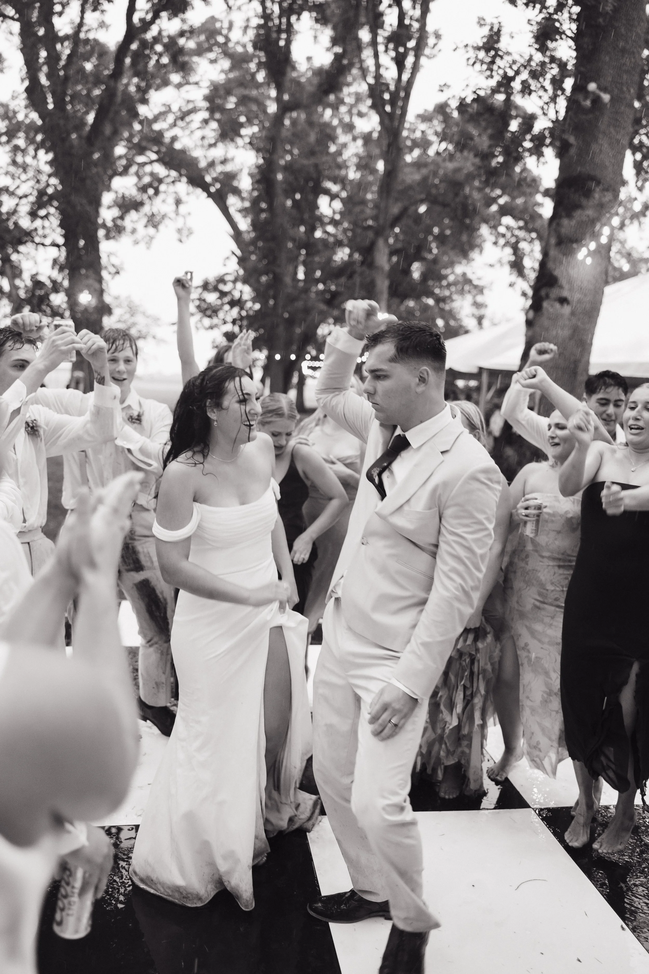 People dancing at an outdoor wedding reception, with a bride in a strapless gown and a groom in a light-colored suit. The scene is lively, with guests raising their hands and enjoying the celebration under trees and string lights.