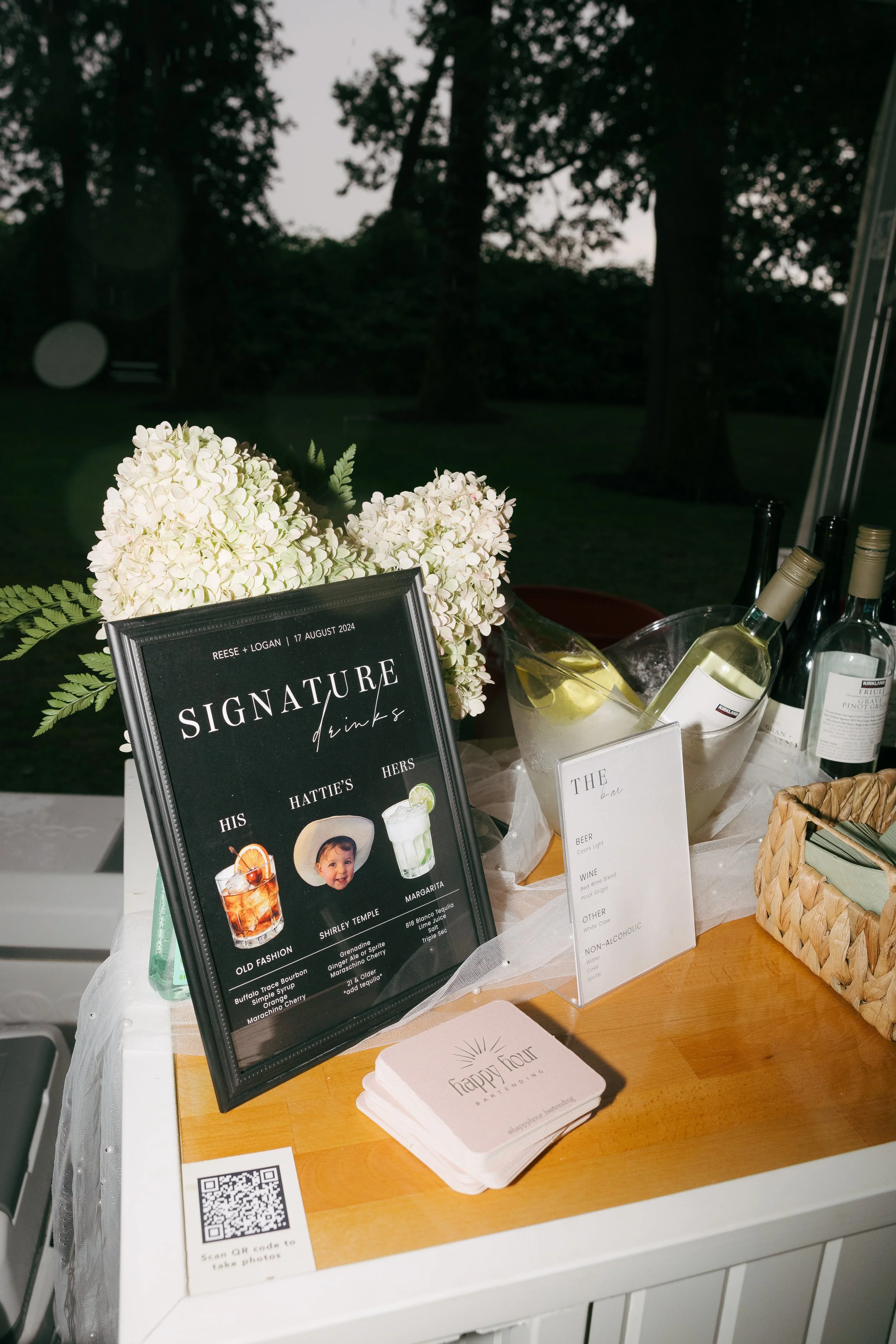 Wedding reception drinks table with floral arrangement, bottled wine and labels, a sign titled 'Signature drinks' with images of cocktails, and a small stack of coasters with a QR code on a white tablecloth.