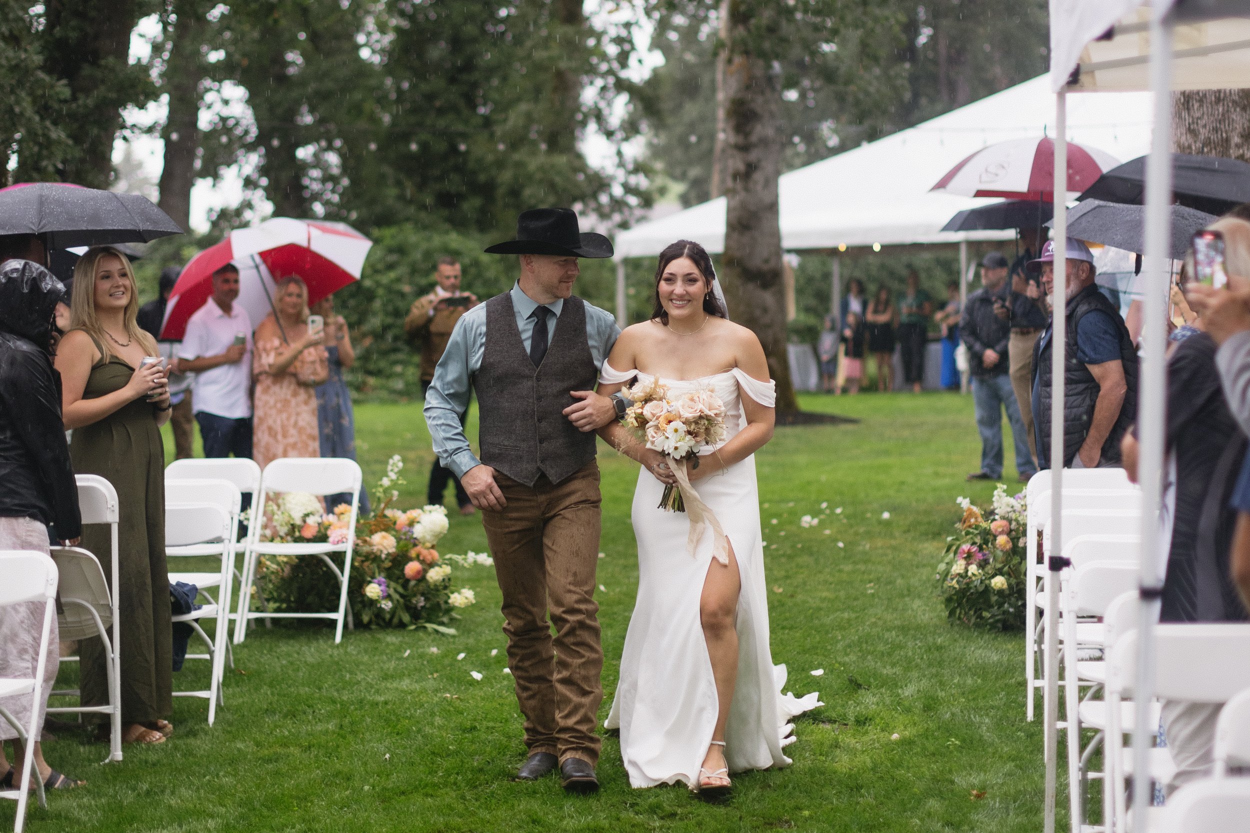 Bride in white wedding dress walking down aisle with man in hat and vest during outdoor wedding ceremony, guests under umbrellas watching in rain.