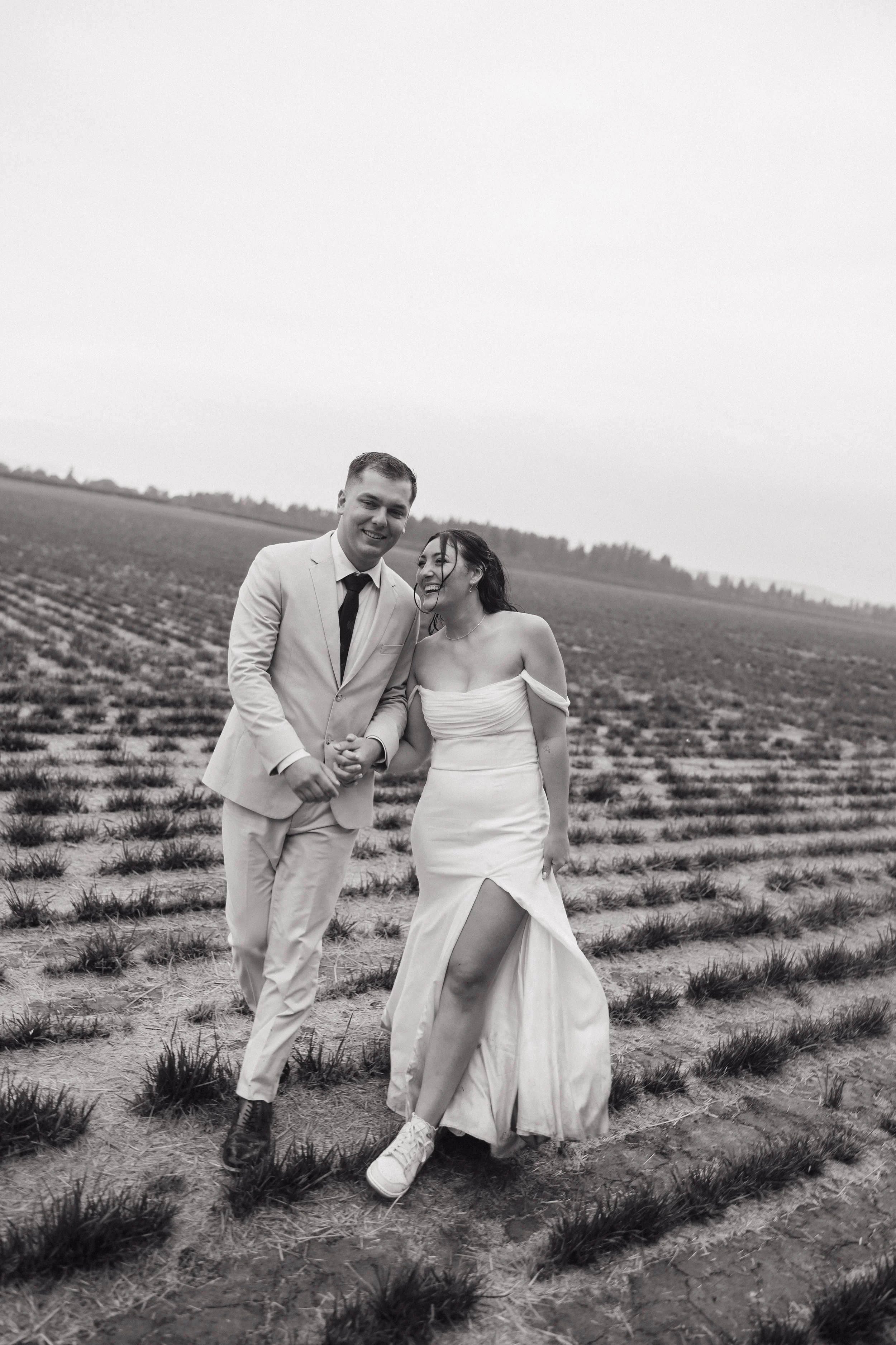A black-and-white photo of a smiling couple walking hand-in-hand in a field, with the woman wearing a strapless dress with a high slit and the man in a light-colored suit and tie.