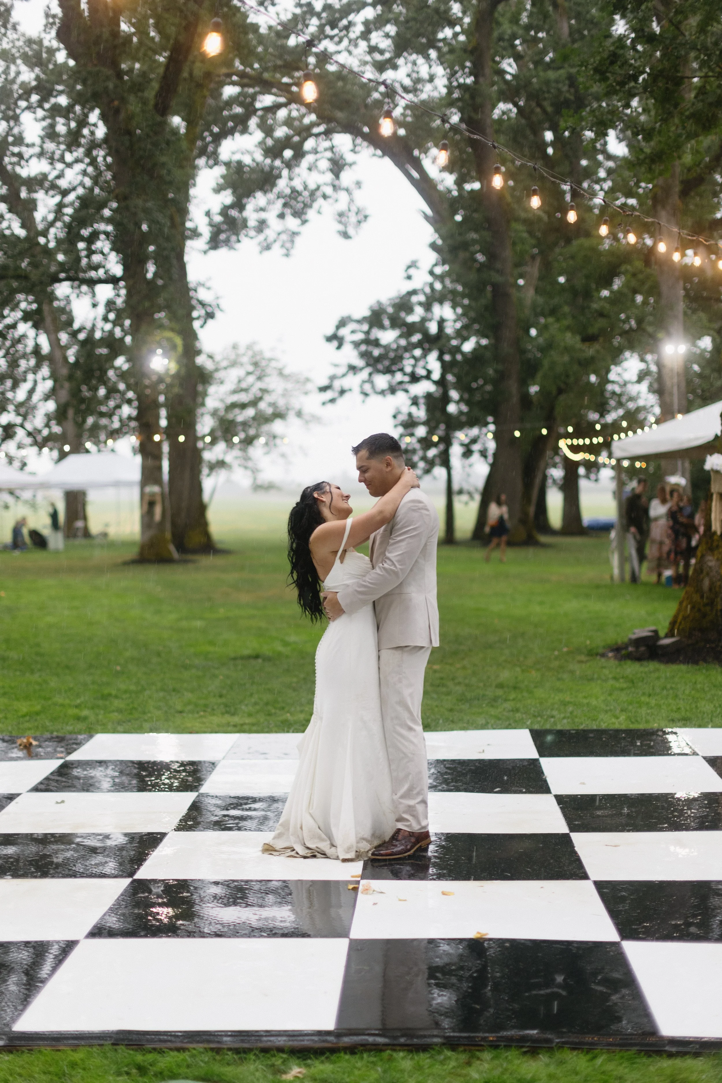 Bride and groom dancing on black and white checkered dance floor outdoors with string lights and trees in the background.