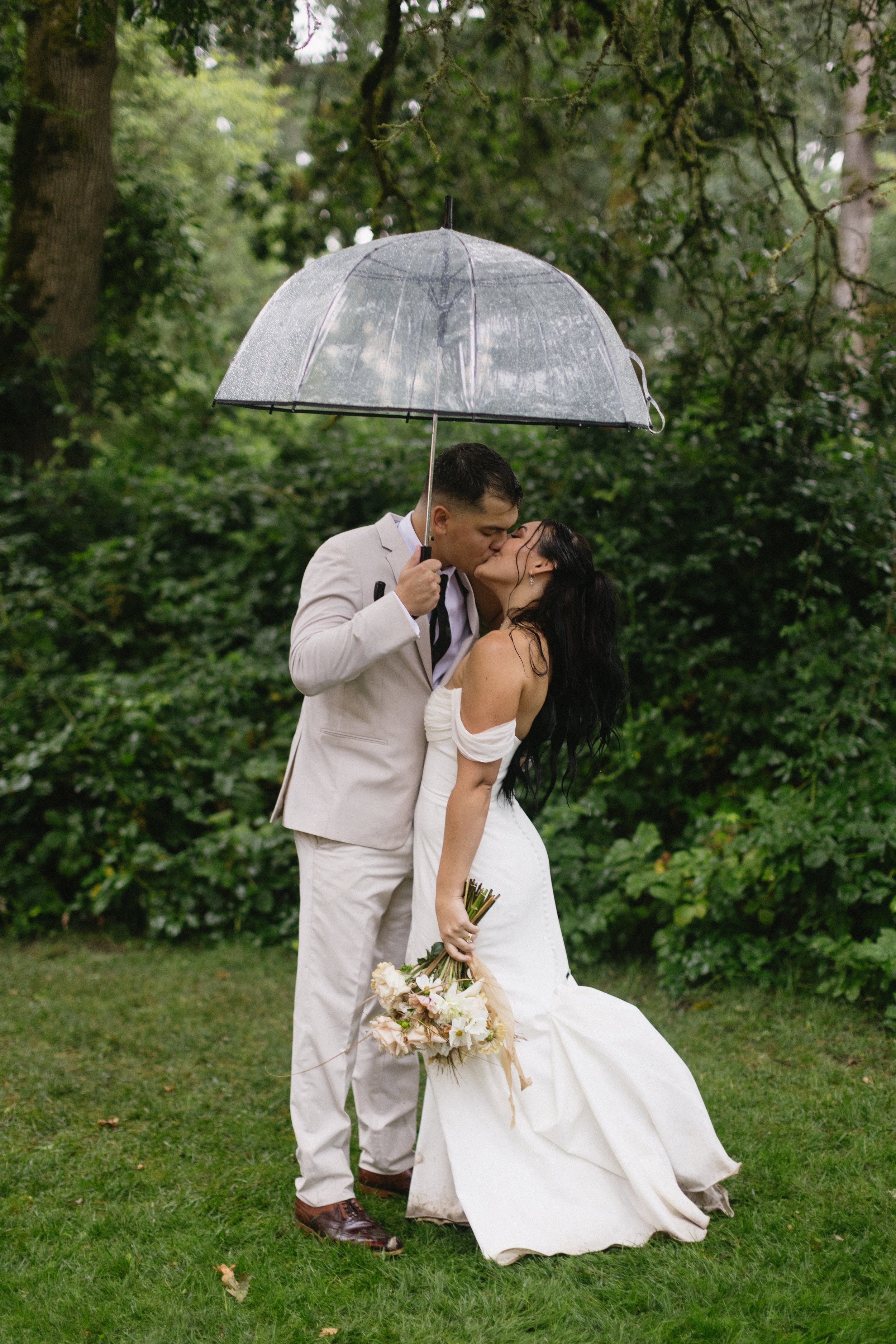 A couple in wedding attire sharing a kiss under an umbrella in a lush, green outdoor setting.