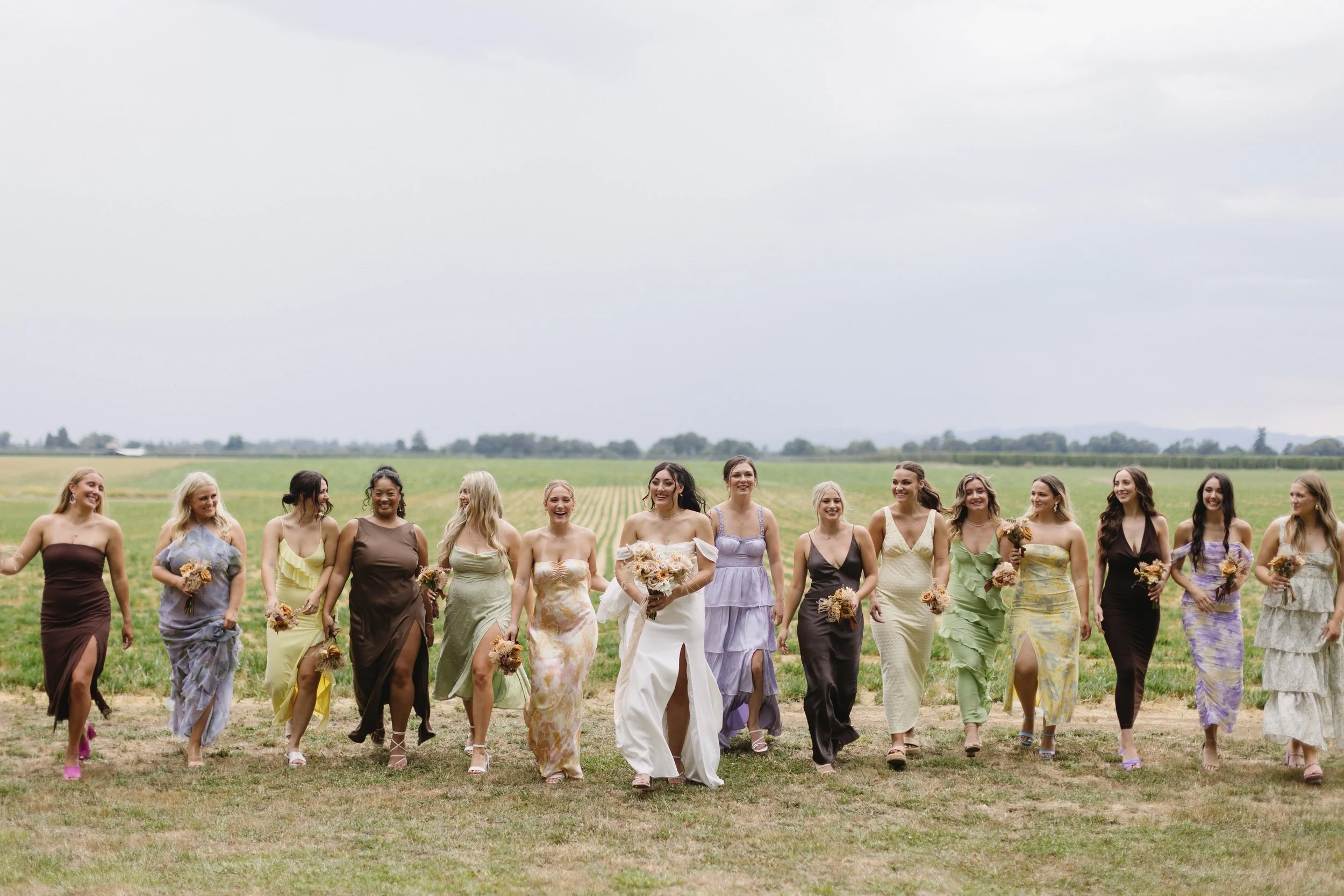 A group of women dressed in colorful dresses walking together outdoors in a rural field, holding bouquets of flowers, smiling and enjoying a sunny day.