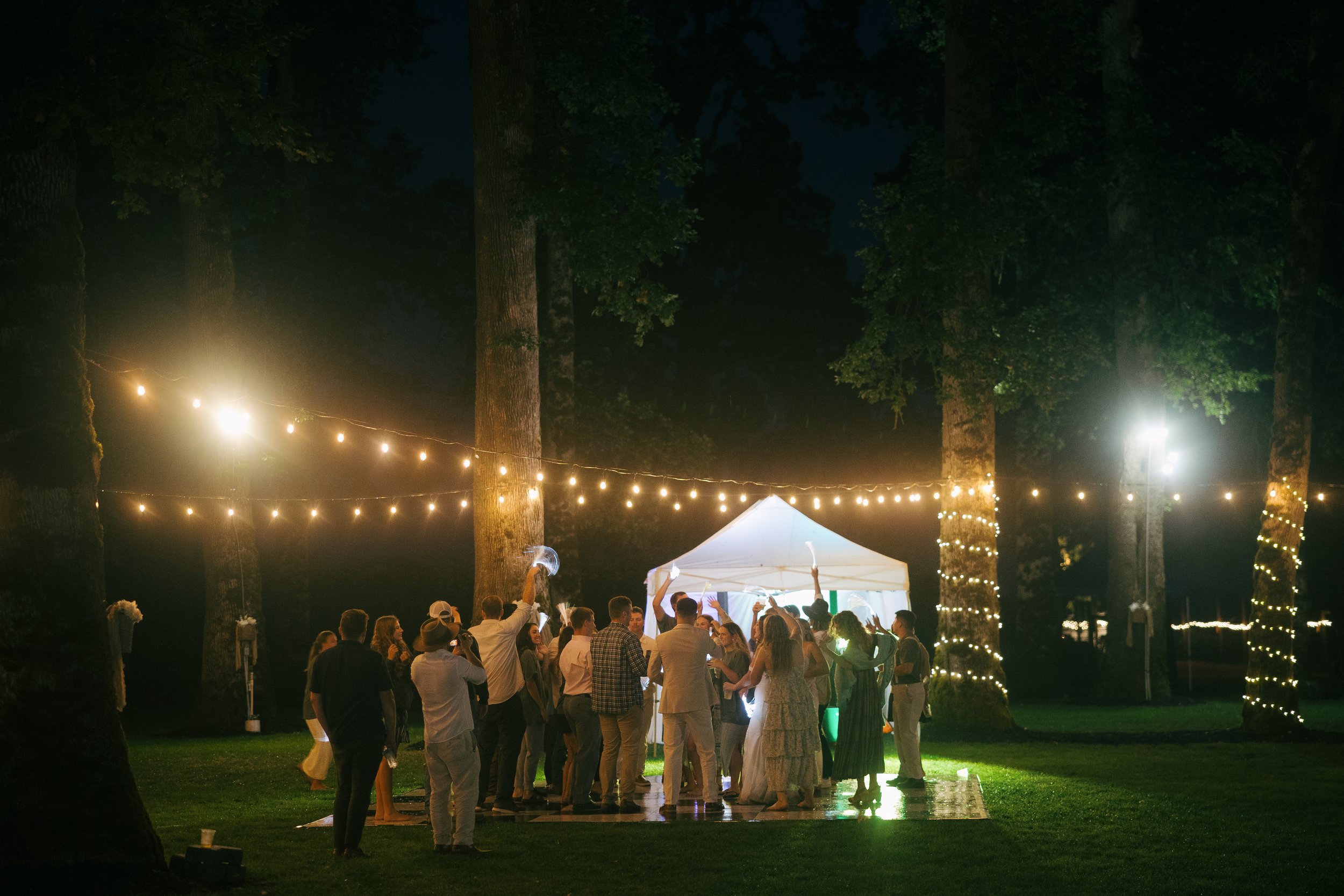 People dancing and celebrating during an outdoor nighttime event under string lights and trees, with a white canopy tent in the background.