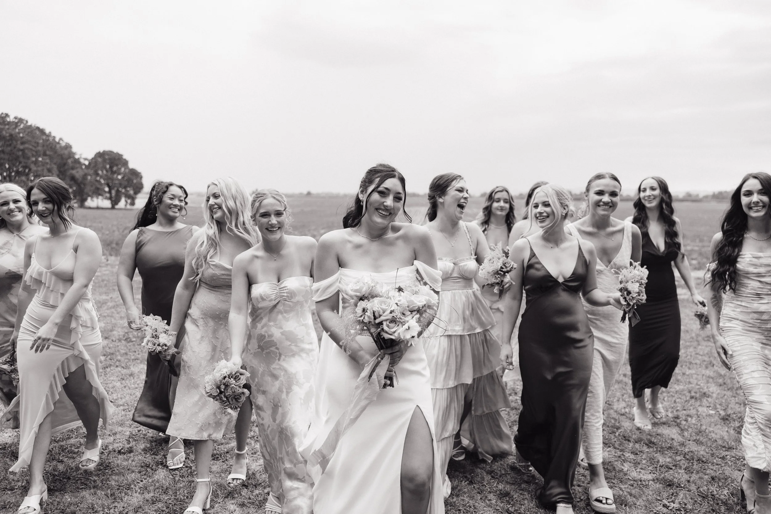 A group of women dressed in formal attire walking outdoors on a grassy field, smiling and laughing, with some holding bouquets of flowers.